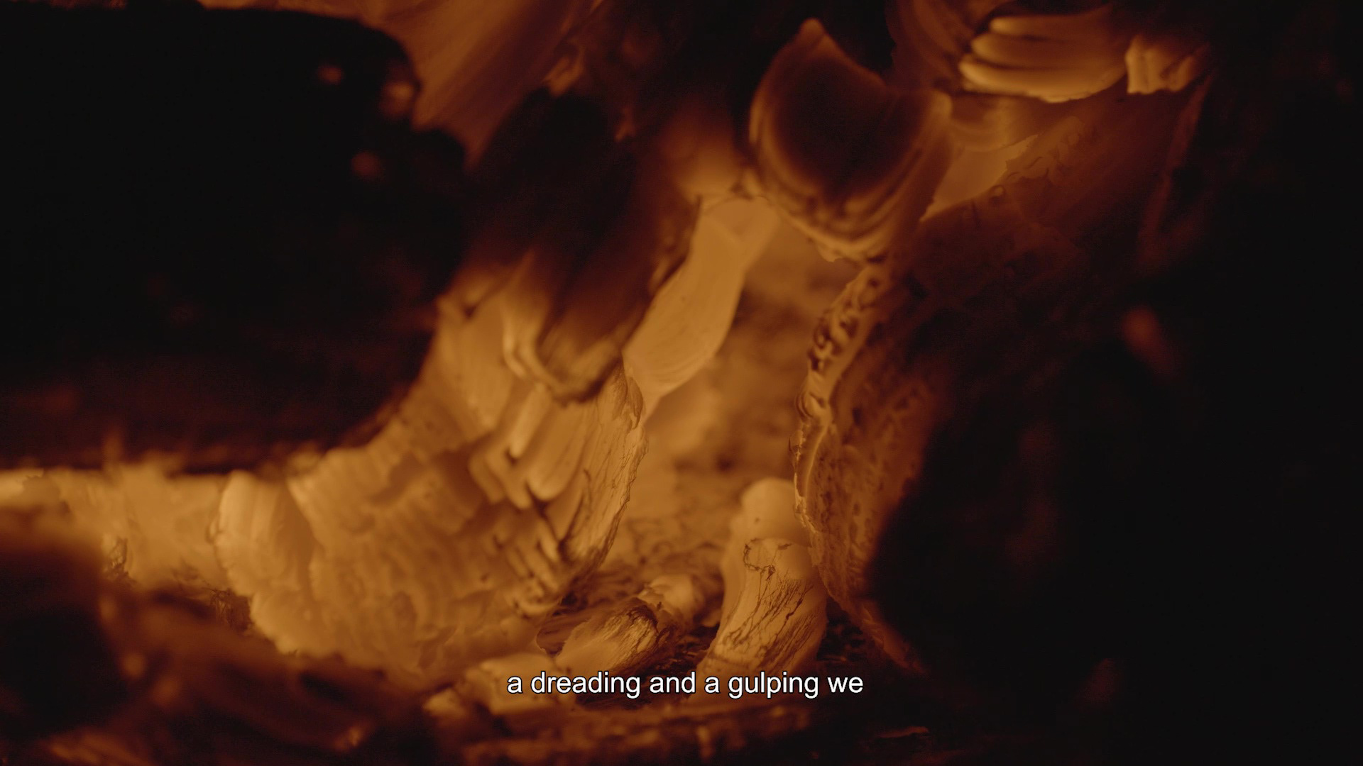 A still from the film Imbolc by Joseph June Bond. A close up of glowing embers that appear to be created from painterly strokes of golden, orange and brown. Captions in white text across the bottom of the screen read ‘a dreading and a gulping we’