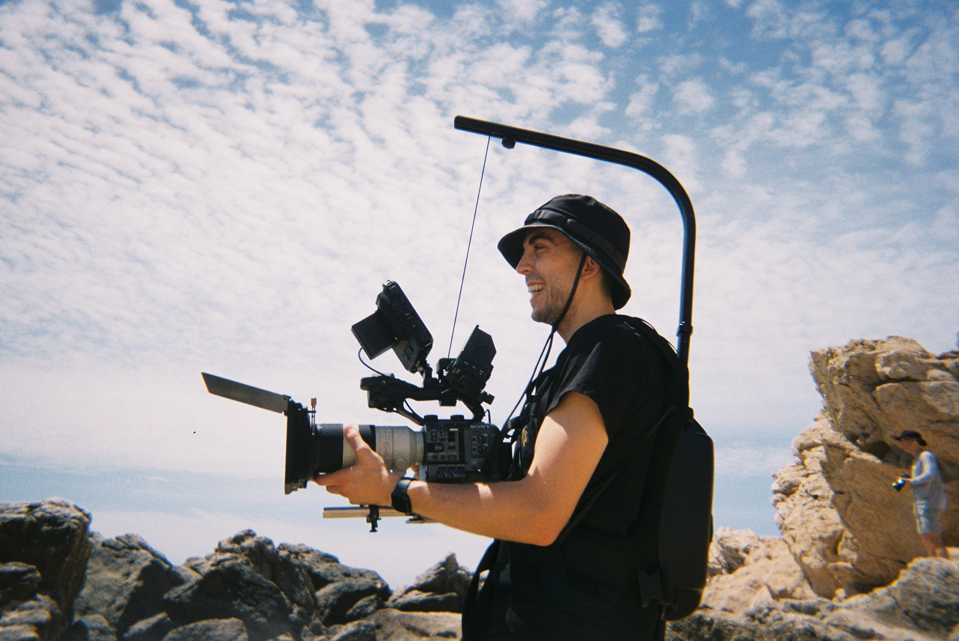 Grant smiling, wearing a camera stabilizer and filming on a sunny beach.