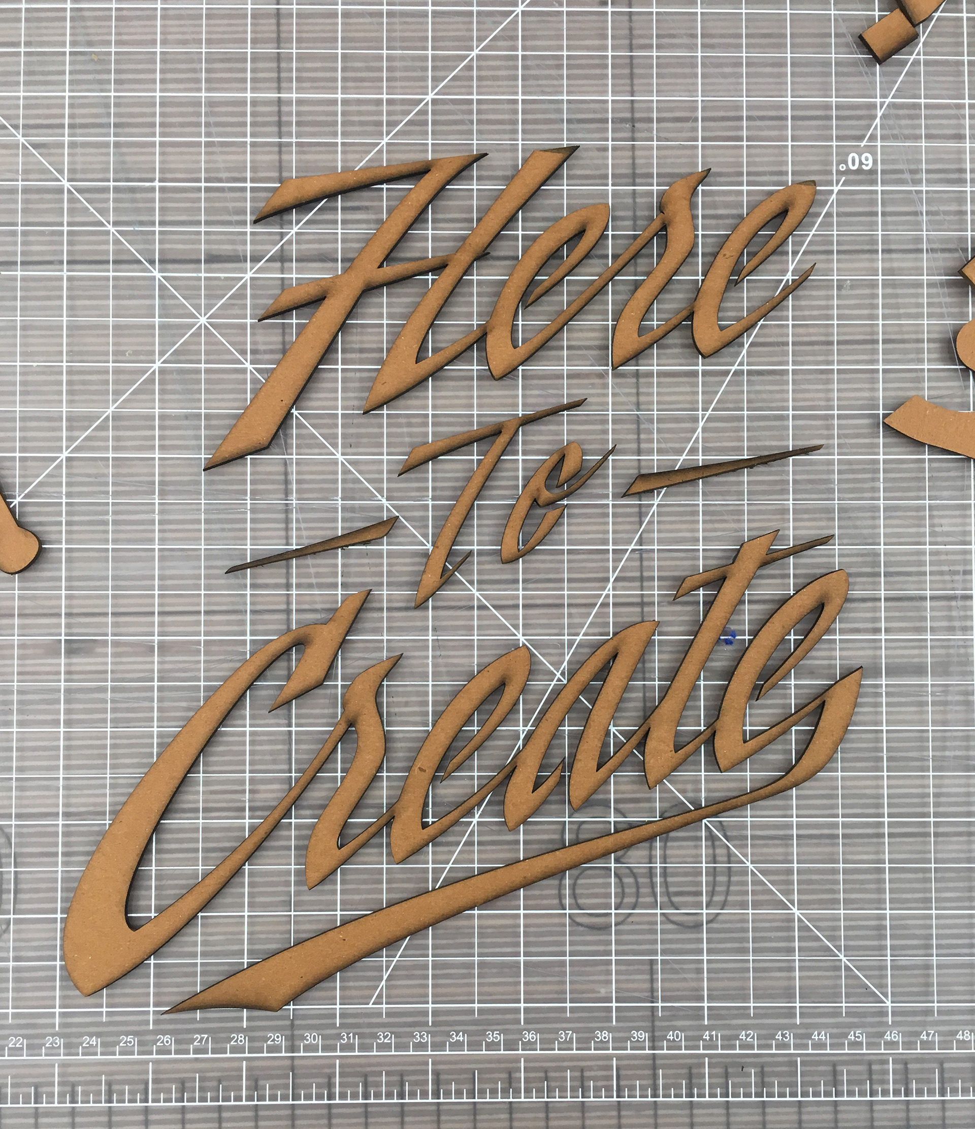 Cardboard cut-out of Here to Create script lettering on an exacto cutting mat.