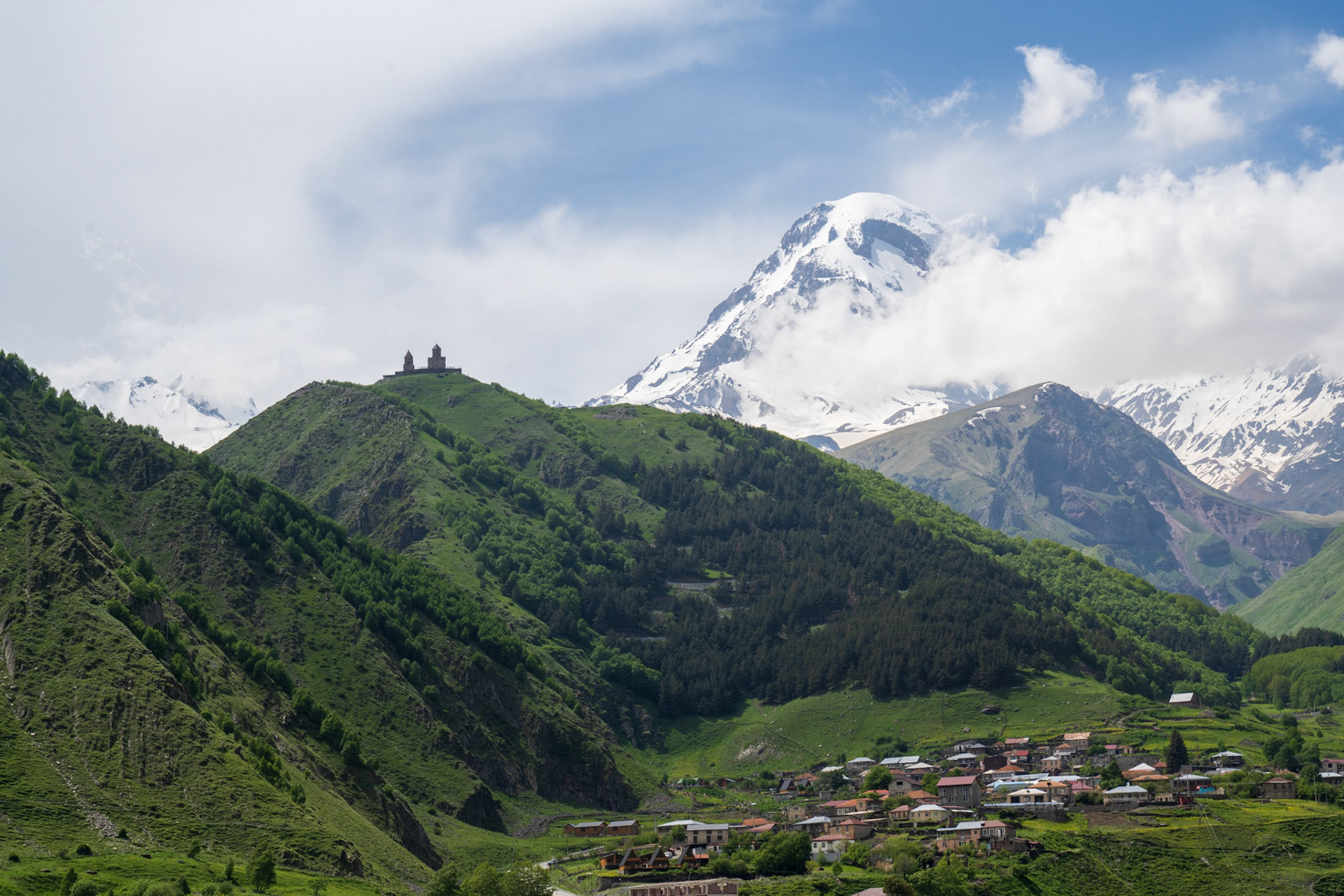 Stepantsminda - The famous hilltop silhouette of Tsminda Sameba Church and the towering snowy cone of Mt Kazbek looking down from the west.