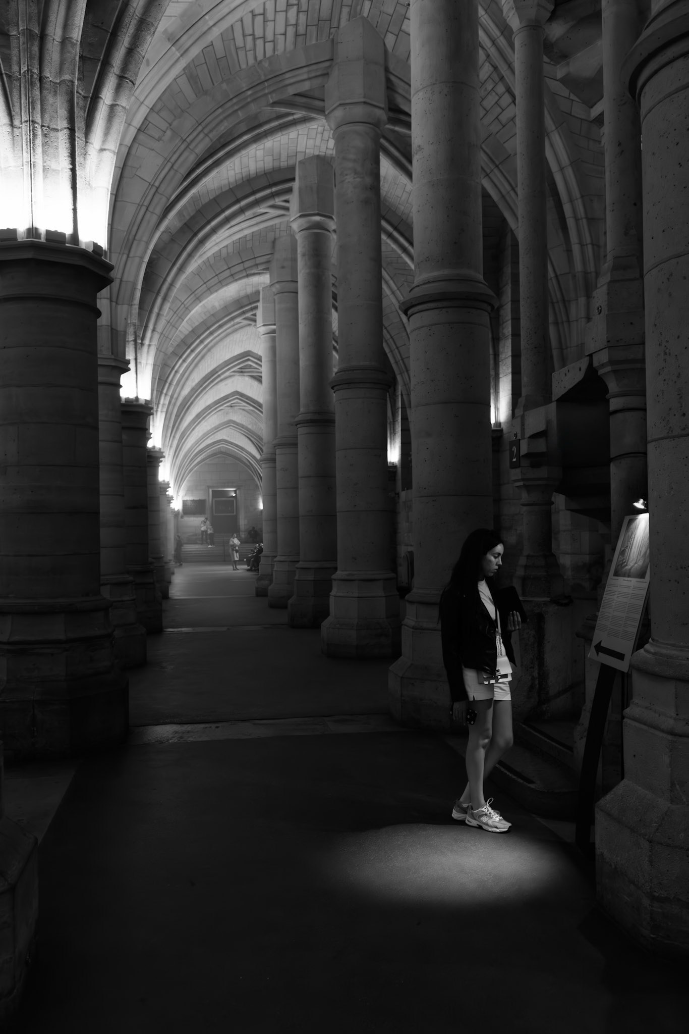 A woman standing in a pool of light in a vaulted gallery at the Conciergerie