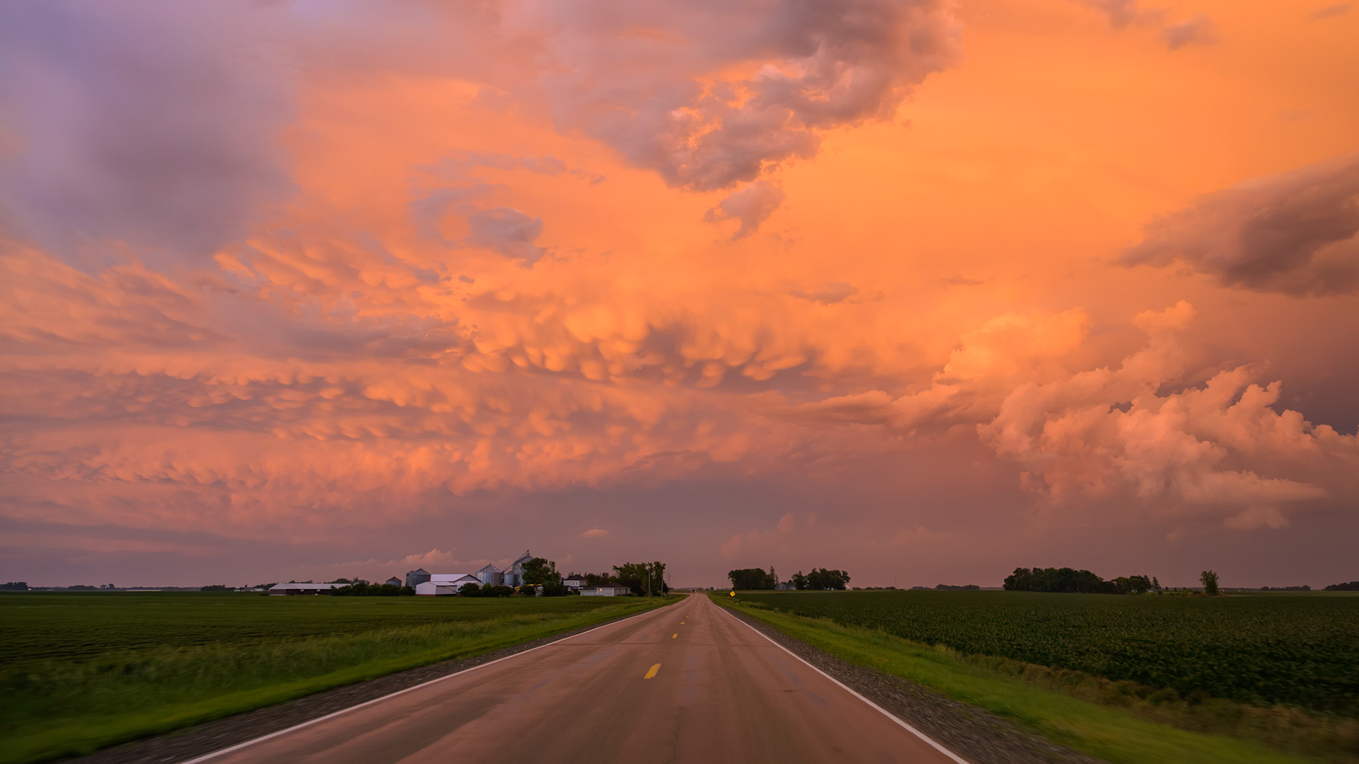 A rural road near Waldorf, Minnesota, provides a stunning view of a post-storm sunset. The sky is filled with dramatic mammatus clouds, illuminated in brilliant orange and purple. These pouch-like formations often appear on the underside of a severe thunderstorm's anvil, indicating turbulent, sinking air—a spectacular sight for storm chasers.