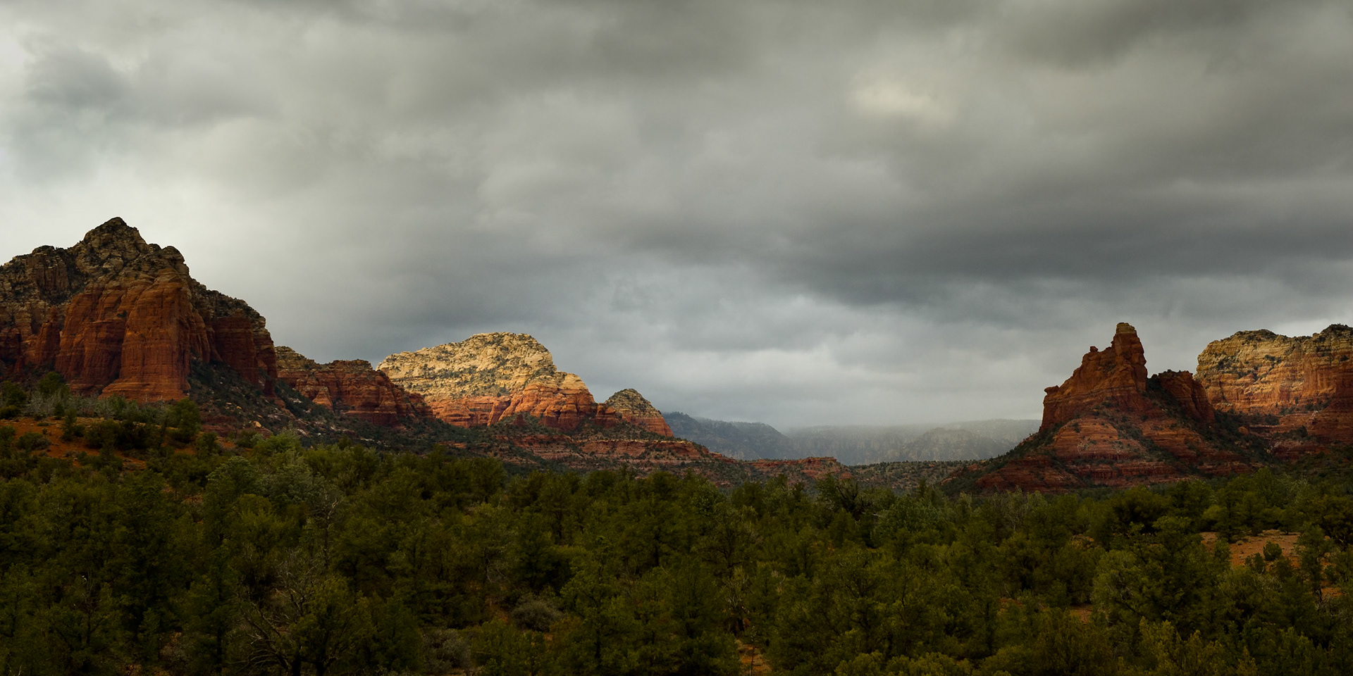 Late afternoon sun breaks through storm clouds to illuminate the iconic red rock buttes of Sedona, Arizona. A rain squall can be seen moving through the distant valley, creating a moment of dramatic, fleeting light over the pinyon-juniper woodland.