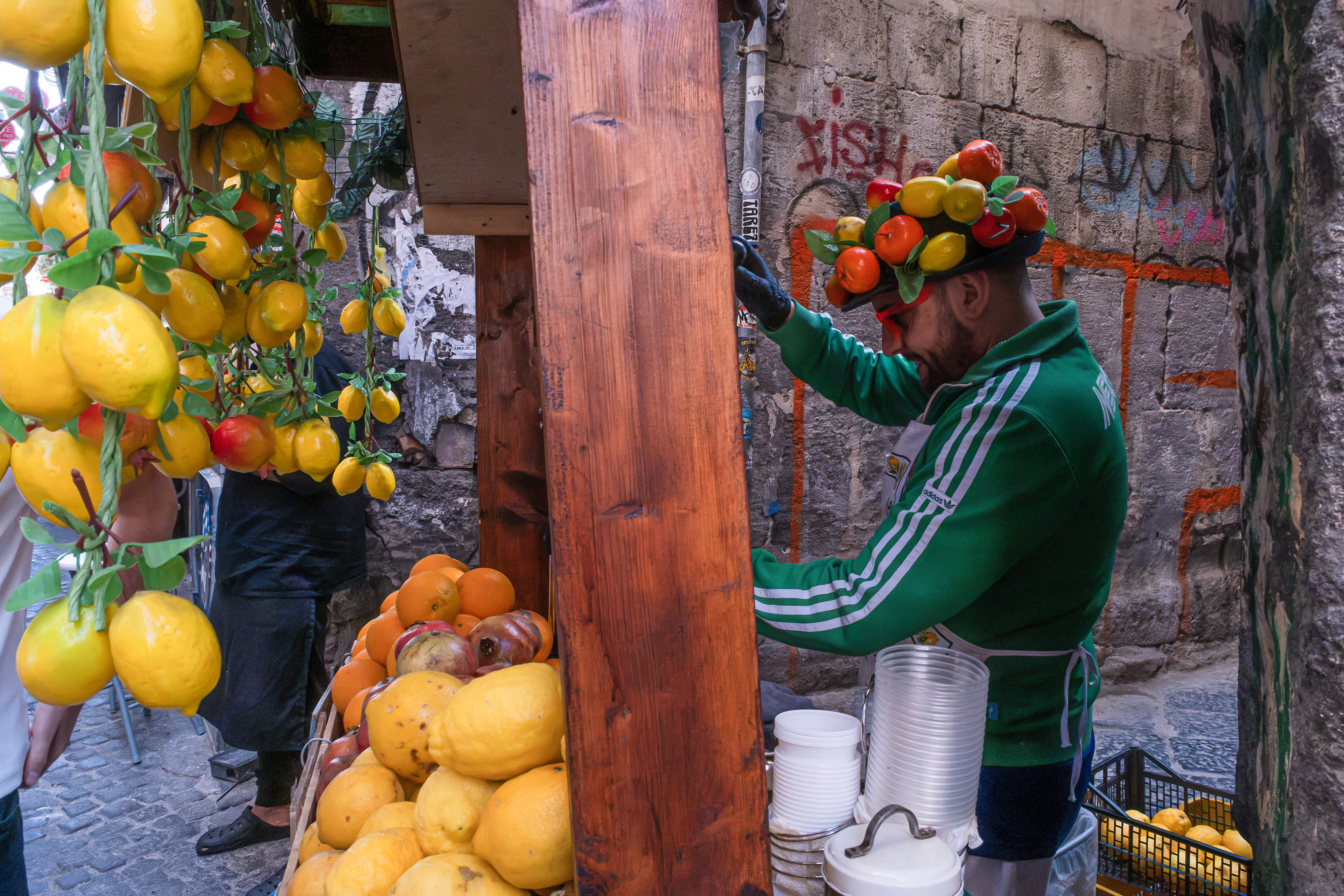 Scena di street photography che ritrae un caratteristico venditore di limonate al lavoro nel suo chiosco di legno in un vicolo storico. L'uomo indossa una giacca sportiva verde e uno stravagante cappello decorato con frutta finta. Sul bancone in legno spiccano una pila di bicchieri pronti per la spremuta, limoni freschi, arance e melograni, incorniciati da un'antica parete in pietra con graffiti.