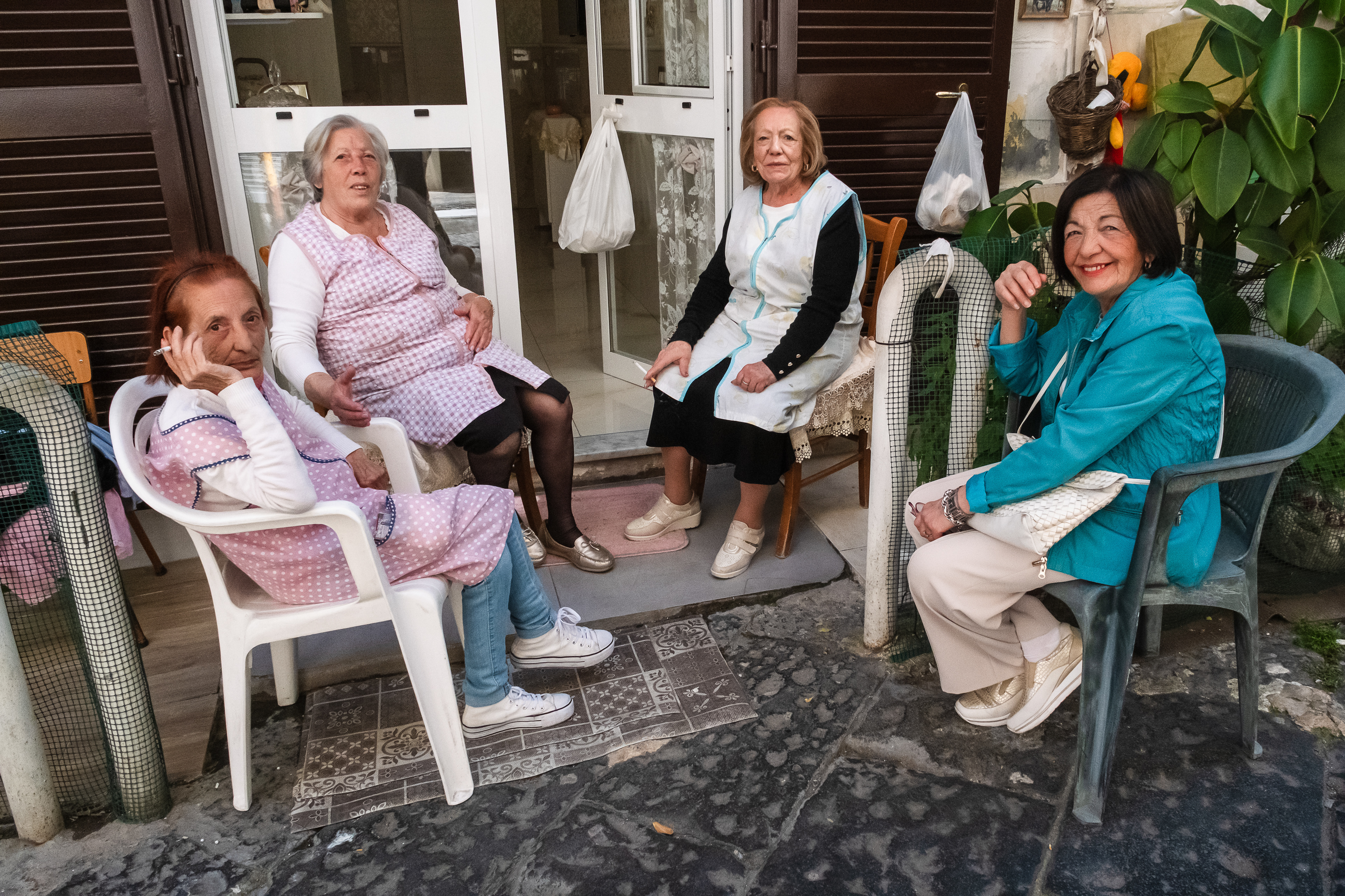 Un'immagine di street photography cattura quattro donne anziane sedute all'aperto in un cortile acciottolato, impegnate in una conversazione informale. Al centro-sinistra, tre donne anziane sono sedute su diverse sedie; due di esse indossano grembiuli a quadri rosa e bianchi sopra maglie bianche a maniche lunghe. La donna a sinistra, con capelli rossi, jeans e sneakers, fuma una sigaretta e guarda verso destra. Dietro di lei, una donna con capelli grigi, anch'essa con un grembiule rosa e sigaretta, guarda verso l'alto. La terza donna anziana, al centro, indossa un grembiule azzurro, una gonna nera e scarpe bianche, sorridendo leggermente. A destra, in primo piano, la quarta donna, con capelli scuri corti, una giacca turchese brillante, pantaloni bianchi e scarpe da ginnastica metallizzate, è seduta su una sedia di plastica verde e sorride direttamente verso la fotocamera. Ha una borsa a tracolla di paillettes bianche. Lo sfondo mostra un edificio con un portone a vetri bianco, tende bianche e persiane di legno scuro chiuse. Sacchetti di plastica bianchi sono appesi a dei ganci sulla porta e sul muro. A sinistra, una recinzione a rete con un piccolo oggetto a forma di sole. A destra, una grande pianta di Ficus elastica (pianta del caucciù) con foglie verdi. L'atmosfera è autentica e sociale, con luce naturale.