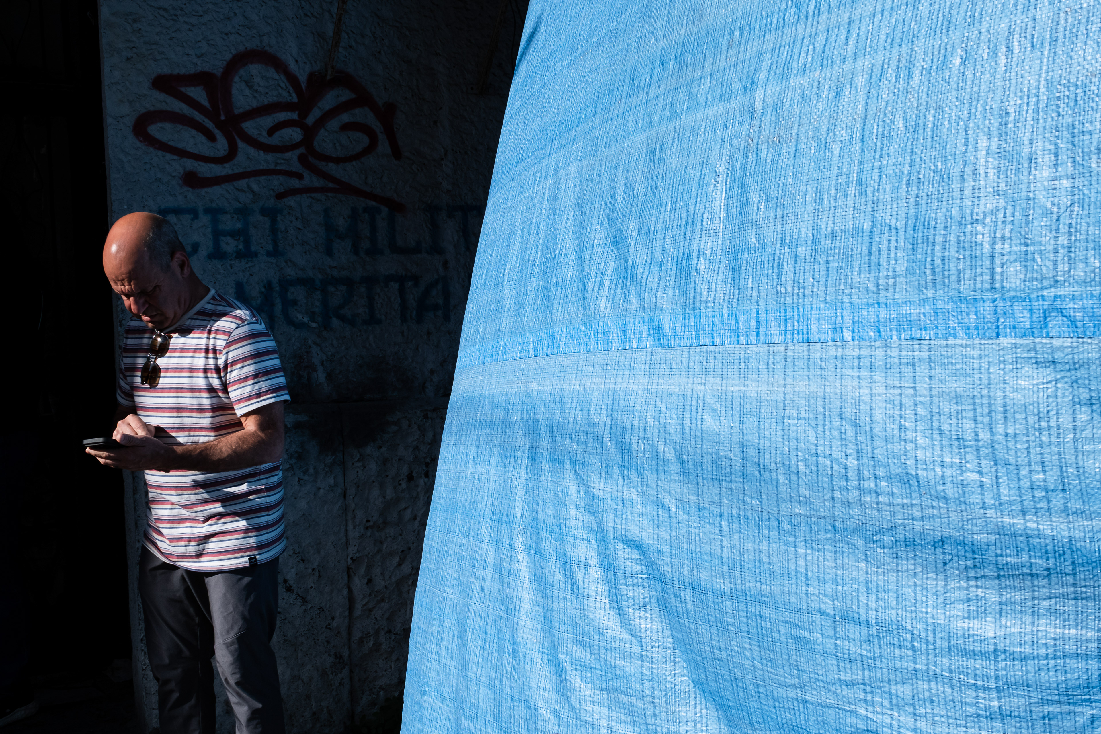 Un uomo calvo in piedi a sinistra, con una maglietta a righe rosse e bianche, occhiali appesi alla scollatura e pantaloni scuri, usa uno smartphone davanti a un muro di pietra scuro coperto da graffiti rossi e una scritta stencil nera che recita "FIGHT HELL AMERICA". La metà destra dell'immagine è occupata da un grande telone di plastica blu brillante intrecciato, illuminato dalla luce solare.