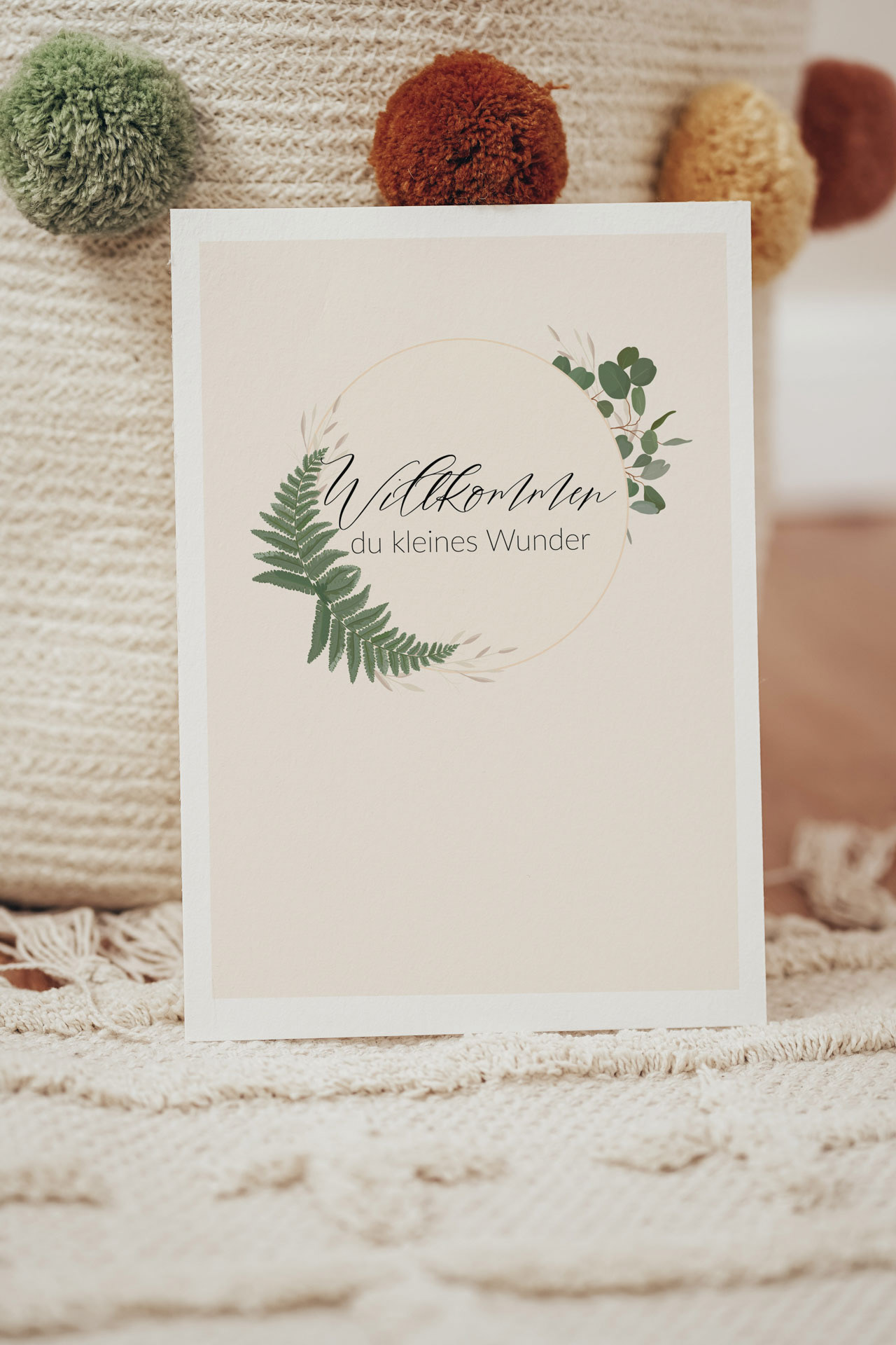 Karte zur Geburt, mit einem goldenen Kreis umgeben von Eukalyptus und Farn, im Zentrum steht der Text: Willkommen du kleines Wunder. Der Hintergrund ist beige und die Pflanzen grün.