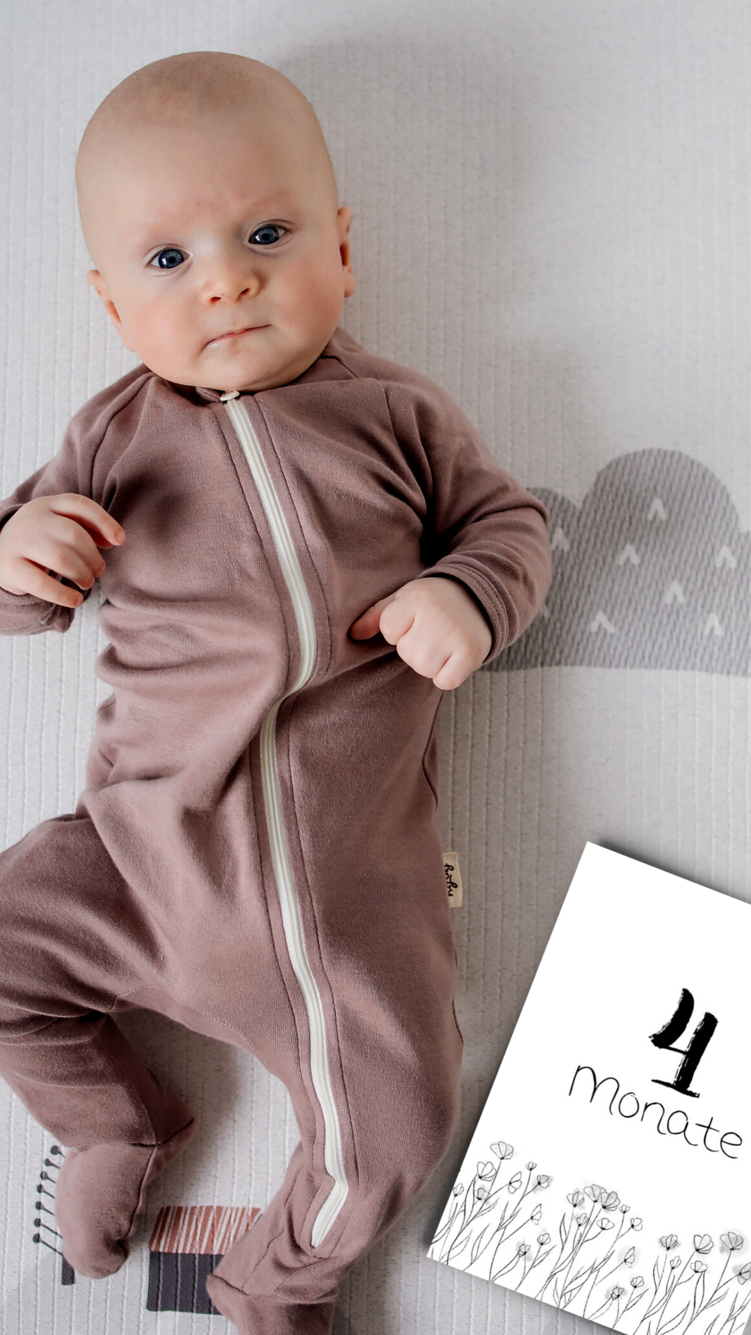 Baby mit altrose Schlafanzug mit Blick in die Kamera. Neben ihm eine Meilensteinkarte mit dem Text: 4 Monate und Zeichnungen von kleinen Blumen in schwarz-weiß mit grauen Aquarell-Kleckse