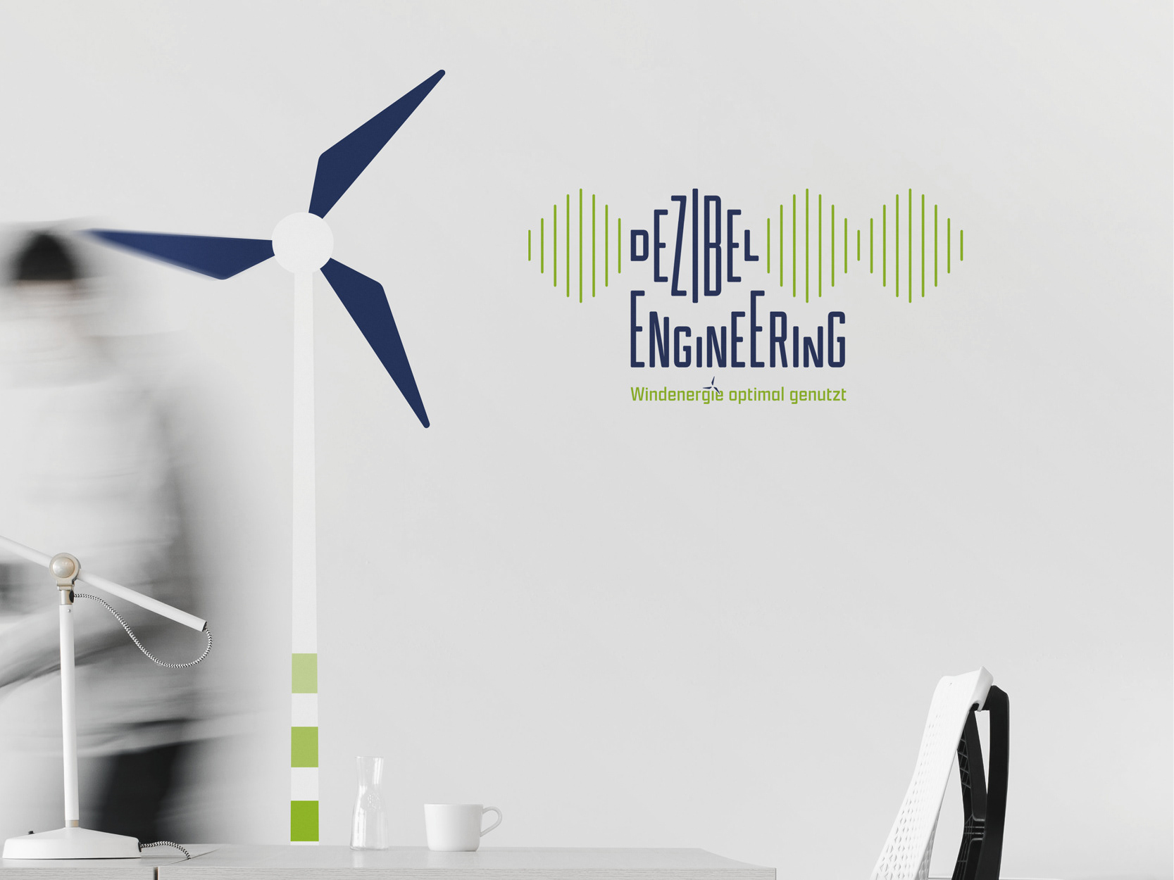 Büroschreibtisch vor einer Wand mit einem Wandtatoo einer Windkraftanlage und dem Logo von "Dezibel Engineering – Windenergie optimal genutzt"