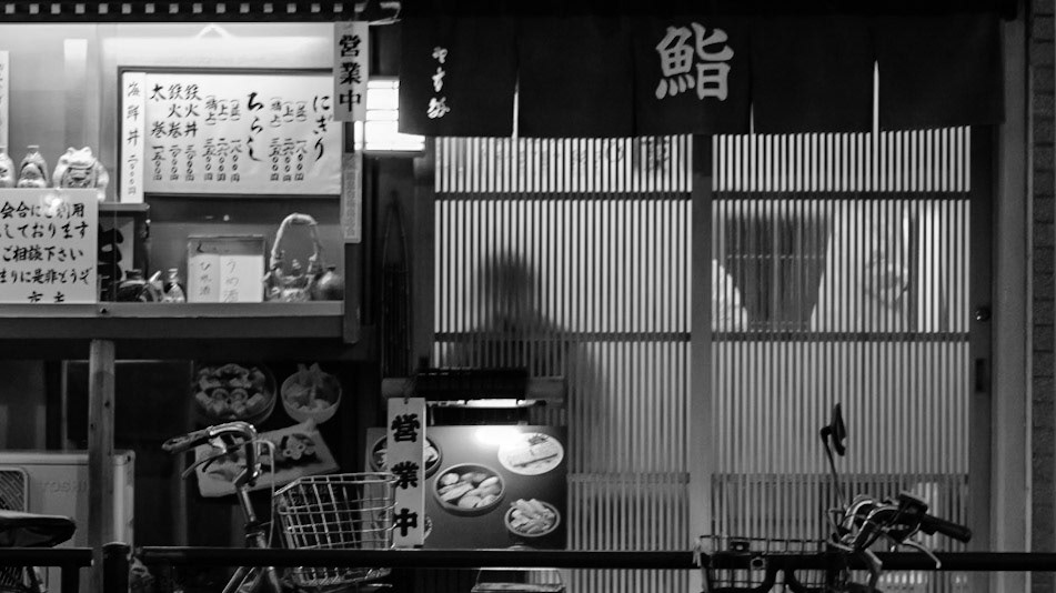 Captured by Ikeji Uju: A person is seen in silhouette through the door of a Sushi resaturant in Taito City, Tokyo, Japan. Shot in Black and White.