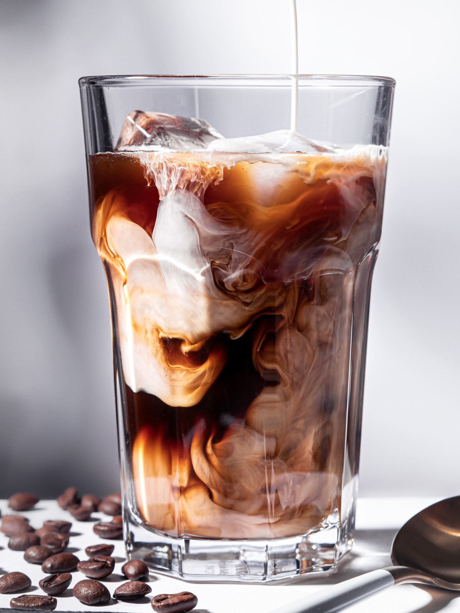 Close up photo of an iced coffee in a glass with white creamer pouring into it and swirling around the glass creating beautiful cascades of coffee and cream in various shapes. Grey background. Glass stands on a white countertop with a white and gold spoon resting next to it, as well as coffee beans spilled around the glass.  