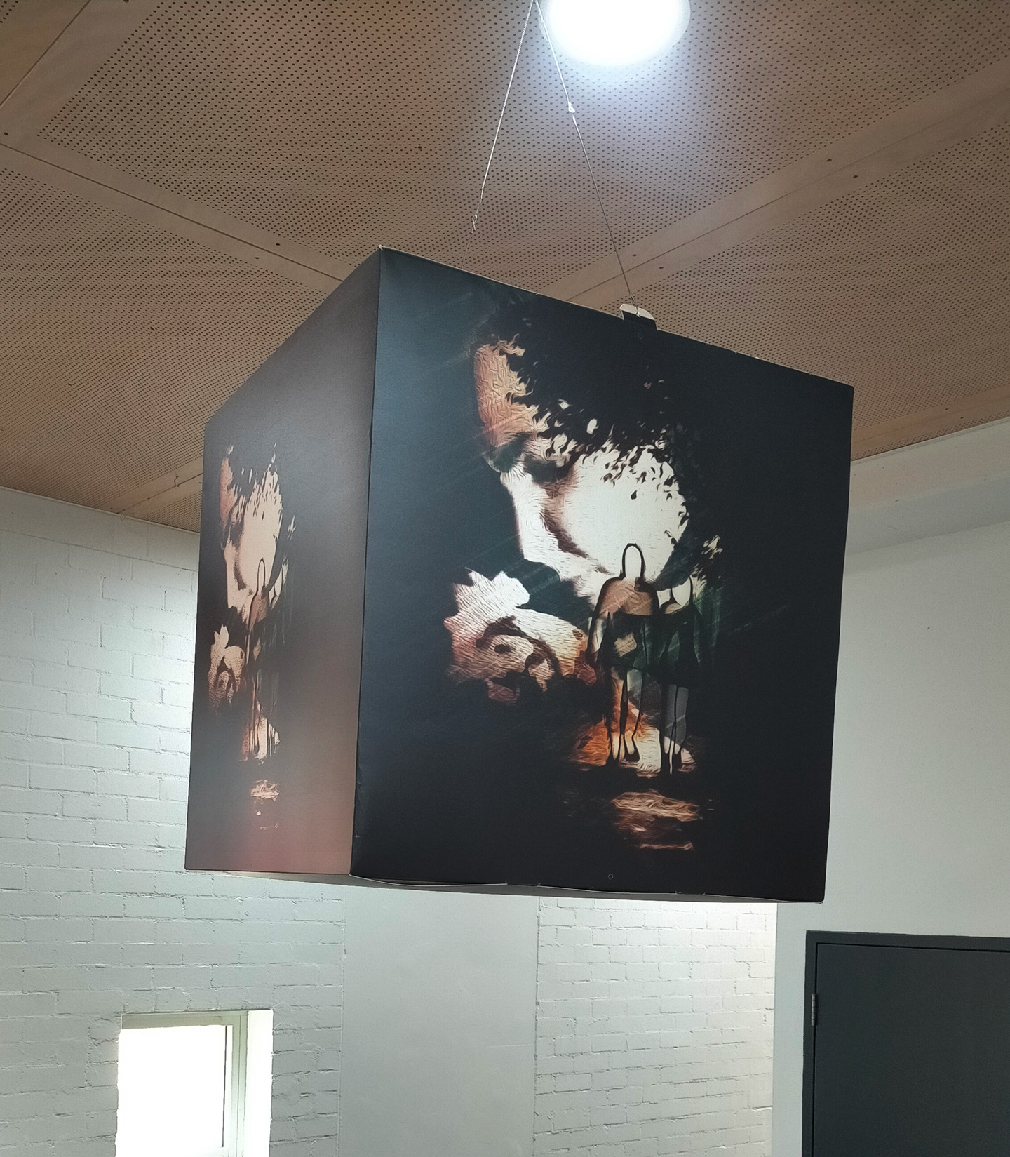 A large photo cube hangs from the ceiling of a large exhibition space. Featured both the front and side of the cube in view is an image of A woman holding a hen blended with a silhouette of a couple walking down a dark foggy street.