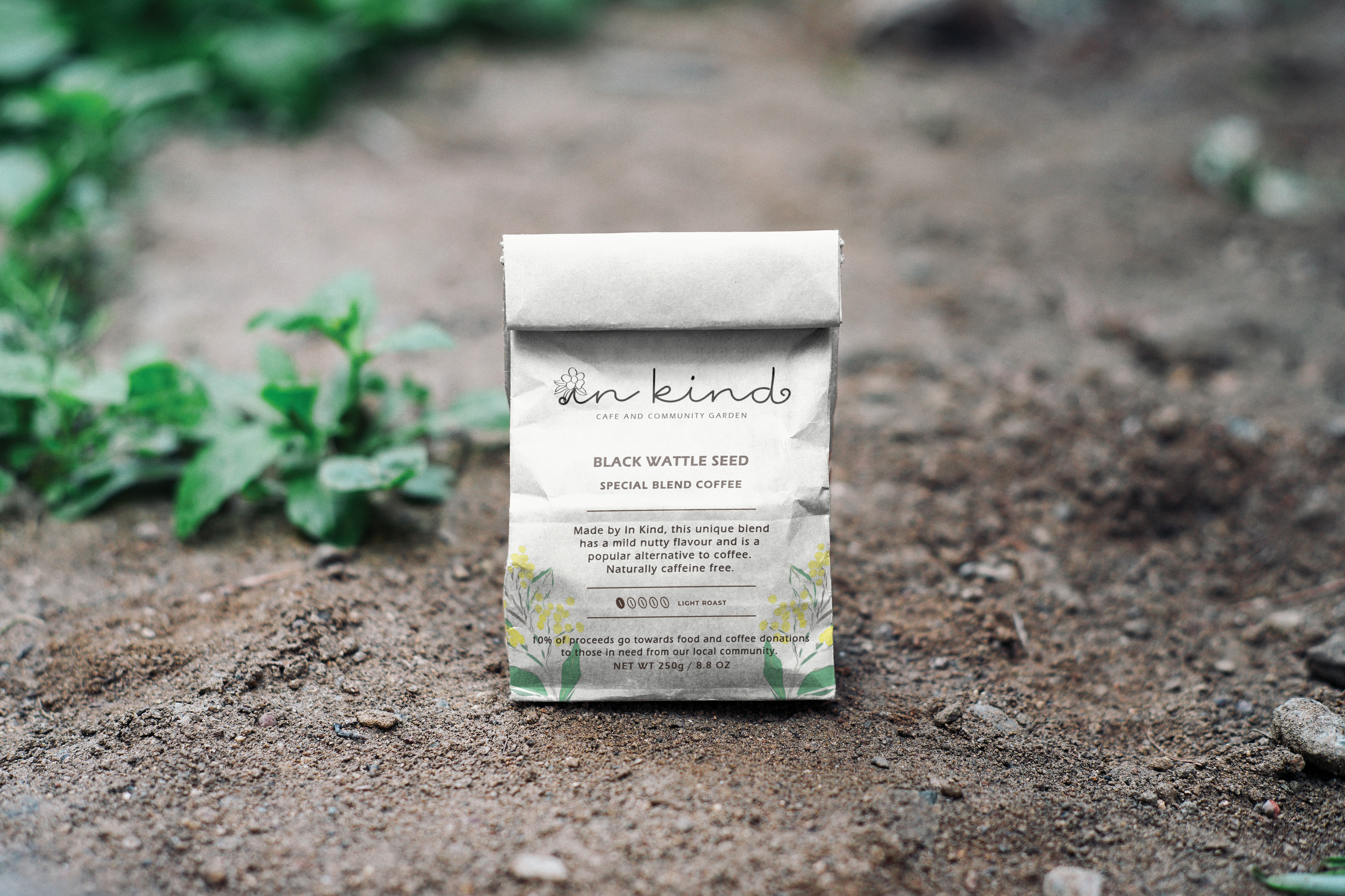 A branding concept mockup for 'In Kind' cafe and community garden featuring a paper bag package for an in-house coffee blend made from black wattle seeds. The packaging displays the custom script logo with a flower replacing the dot on the 'i', and features the hand-drawn wattle motif consistent with the brand identity. The pack includes a promise that 10% of proceeds go towards food and coffee for those in need in the local community