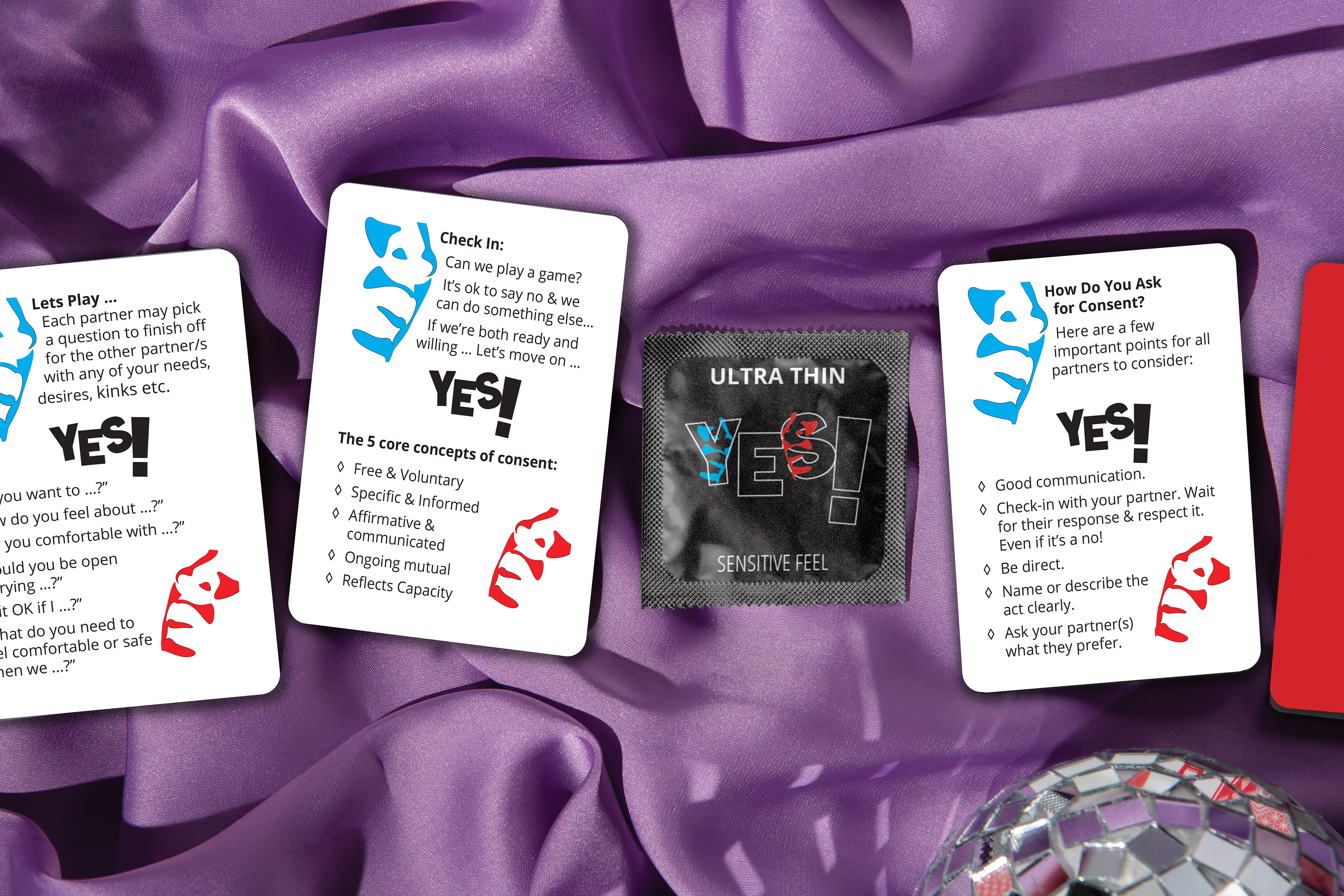A branding and package concept mockup for a condom brand  promoting enthusiastic consent featuring a black condom packet with logo branding on the front. The packet sits amongst three playing cards that describe a game designed to teach and promote conversations around consent. All items rest on a shiny purple material with a mirror ball positioned to the bottom right of the image. The logo is the word YES! with a blue outlined faced imposed on the Y and a Red Outline face on the S. 