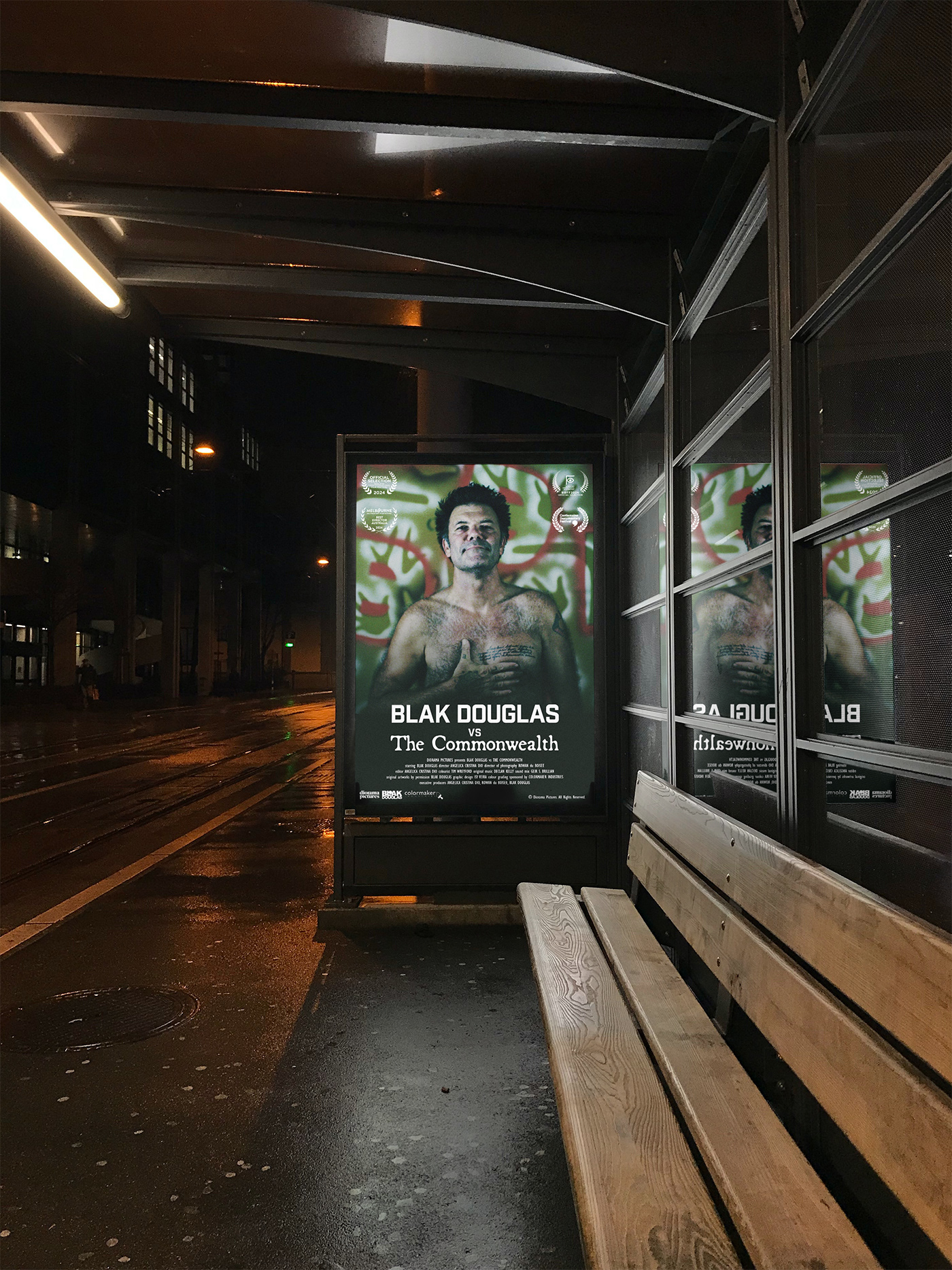 A movie poster mockup displayed in a bus shelter at night for the film 'Blak Douglas Vs The Commonwealth'. The poster features Blak Douglas staring directly at the camera with his right hand placed on his chest, standing against a green wall painted with motifs. In the foreground is the movie title. credits and sponsors in white type over a dark gradient overlay. There are also 4 laurels from Documentary festivals and appearances. 