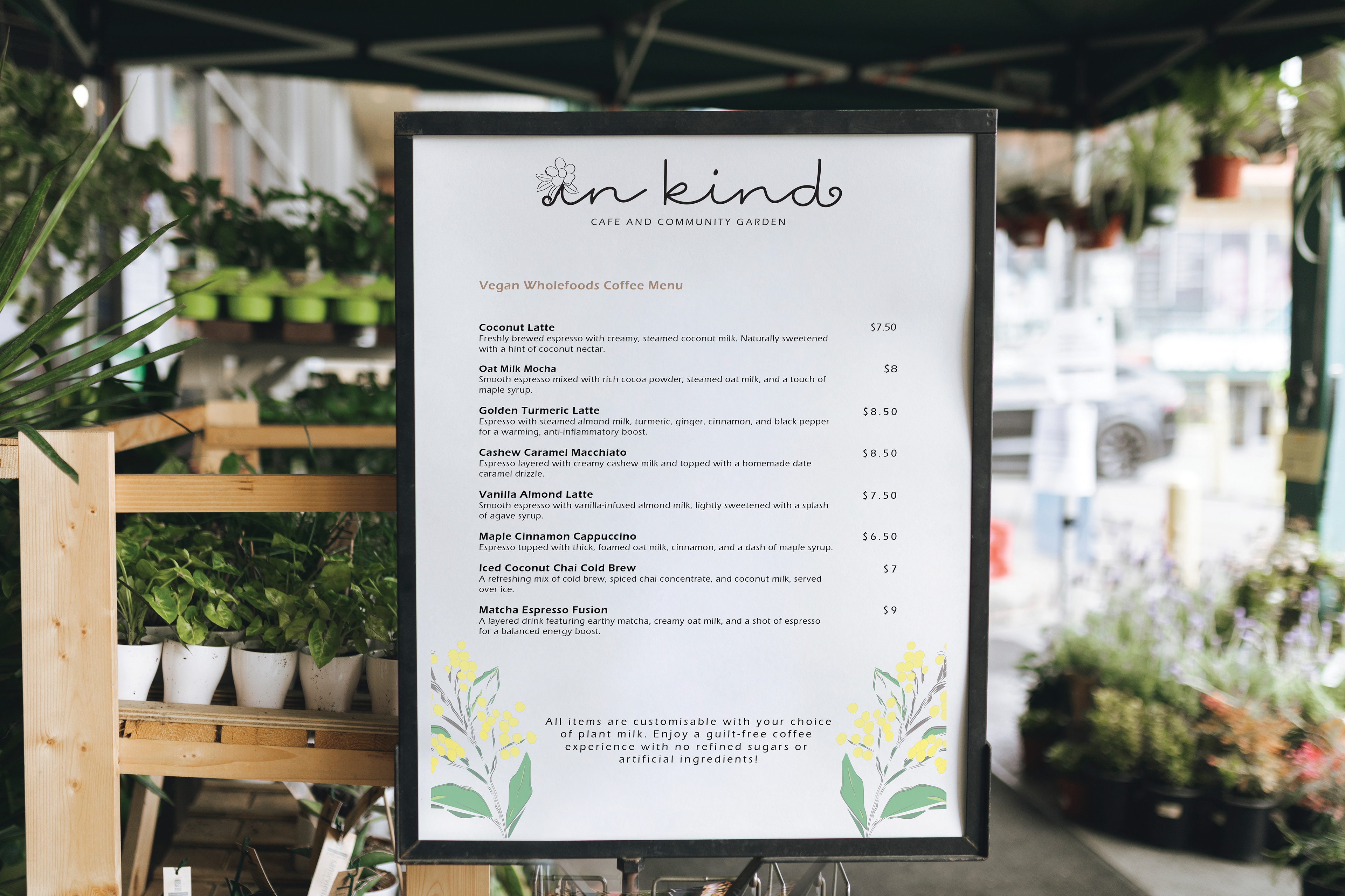 A branding concept mockup for 'In Kind' cafe and community garden featuring a framed sign on a stand with a community garden in the background. The white sign displays the vegan wholefoods coffee menu offered at the cafe, with the custom script logo in black type featuring a flower replacing the dot on the 'i' and the hand-drawn wattle motif consistent with the brand identity
