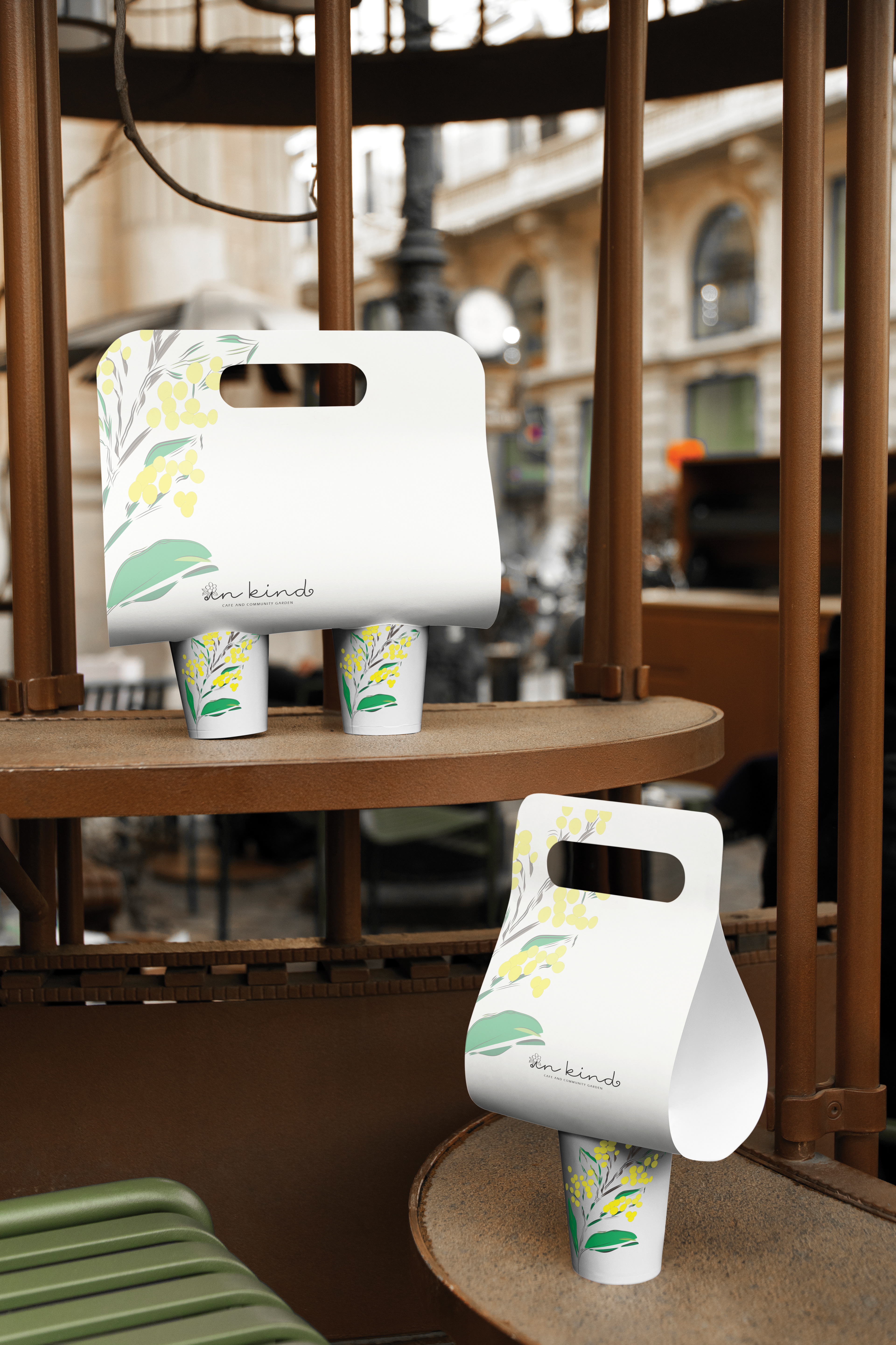 A branding concept mockup for 'In Kind' cafe and community garden featuring takeaway coffee carry trays. The trays display the custom script logo with a flower replacing the dot on the 'i', and the hand-drawn wattle motif consistent with the brand identity
