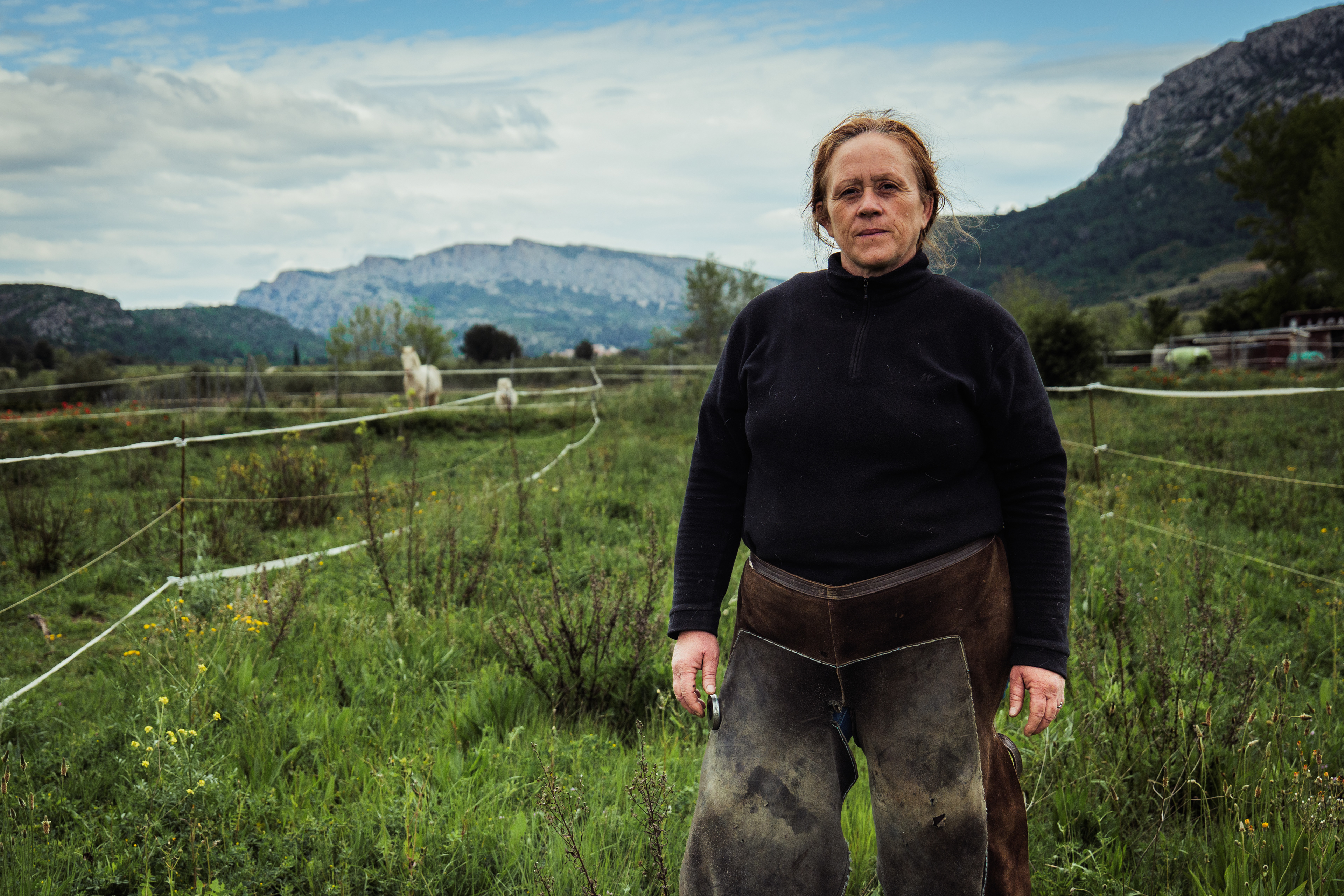 Maud, maréchale-ferrante, pose devant ses terres à Tautavel dans le sud de la France, en avril 2025. En arrière-plan, son Shetland et son Quarter Horse sont dans un pré. Maude est l'une des seules femmes maréchale-ferrantes de France. Dans les Pyrénées-Orientales, elle exerce un métier ancestral à la croisée de l’artisanat, du soin vétérinaire et de l’éthologie et qui est aujourd'hui menacé par la montée de pratiques non qualifiées.