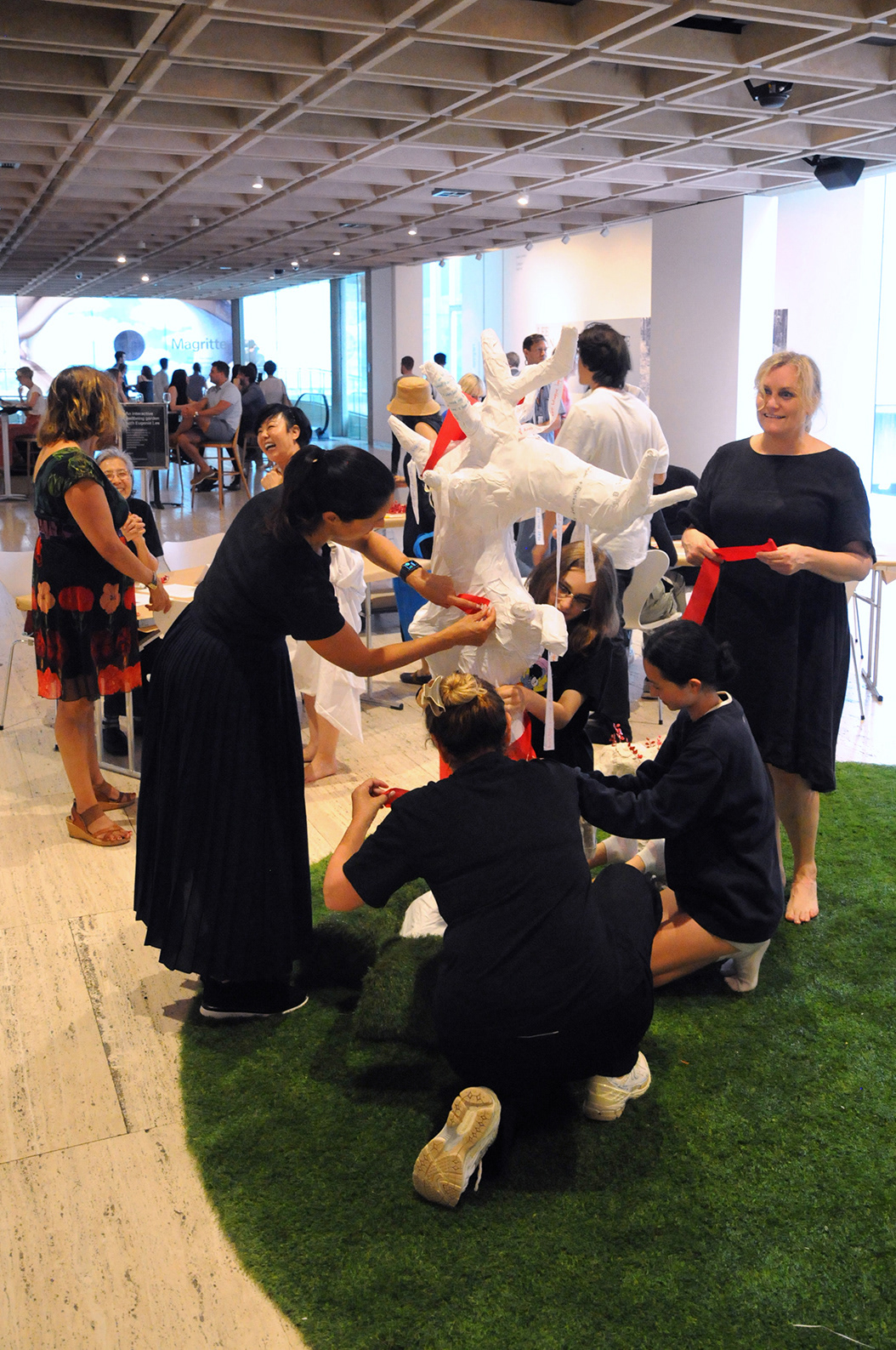 Five people dressed in black wrapping red ribbons around a white tree trunk on a bed of green grass. Other people stand in the background chatting.