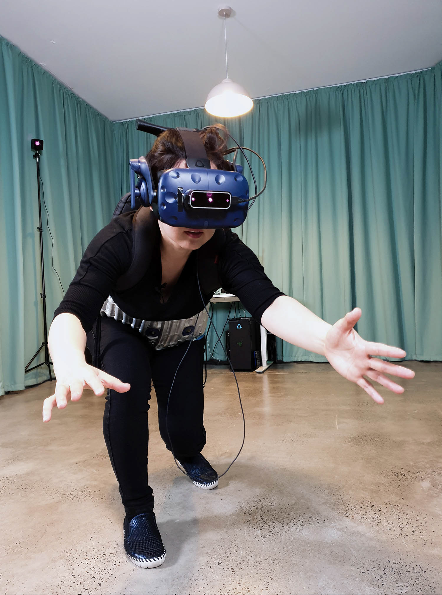 a woman wearing black shirt and pants leaning forward with her hands out-stretched towards the camera. She wears blue virtual reality goggles and large silver belt against a green curtained backdrop.
