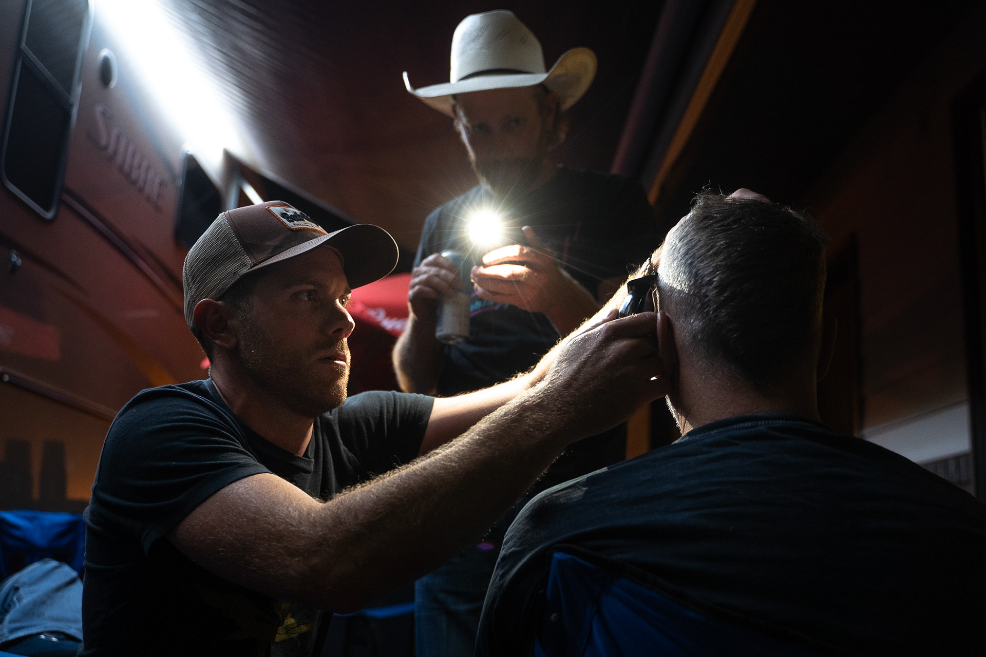 Bryan Loweree, left, cuts lines into the hair of Jay Lee (right) of the Chancey Williams Band as Chancey Williams shines the light during the fourth day of the 81st Sturgis Motorcycle Rally in Sturgis, South Dakota. 08.09.21.