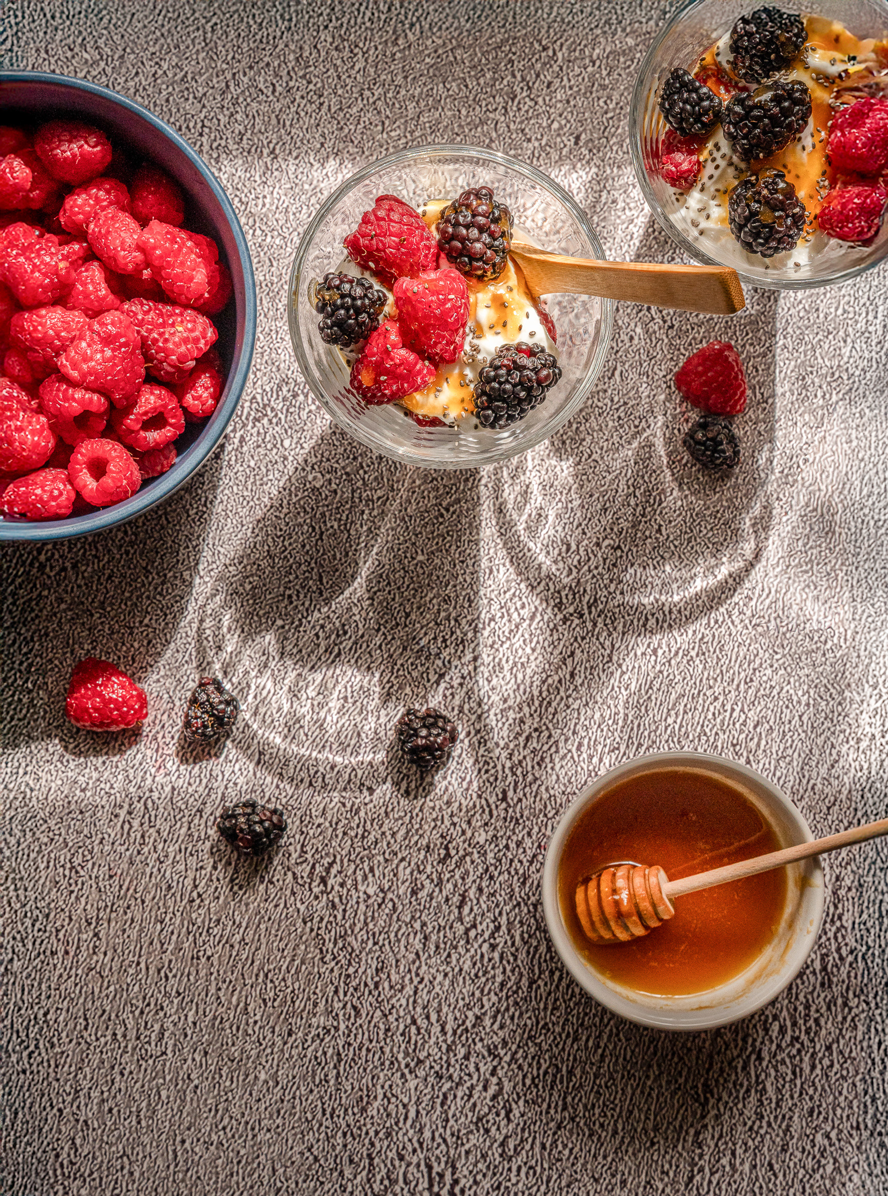 Top-down flatlay photography of fresh red raspberries in a dark bowl on a textured neutral background with harsh artistic shadows