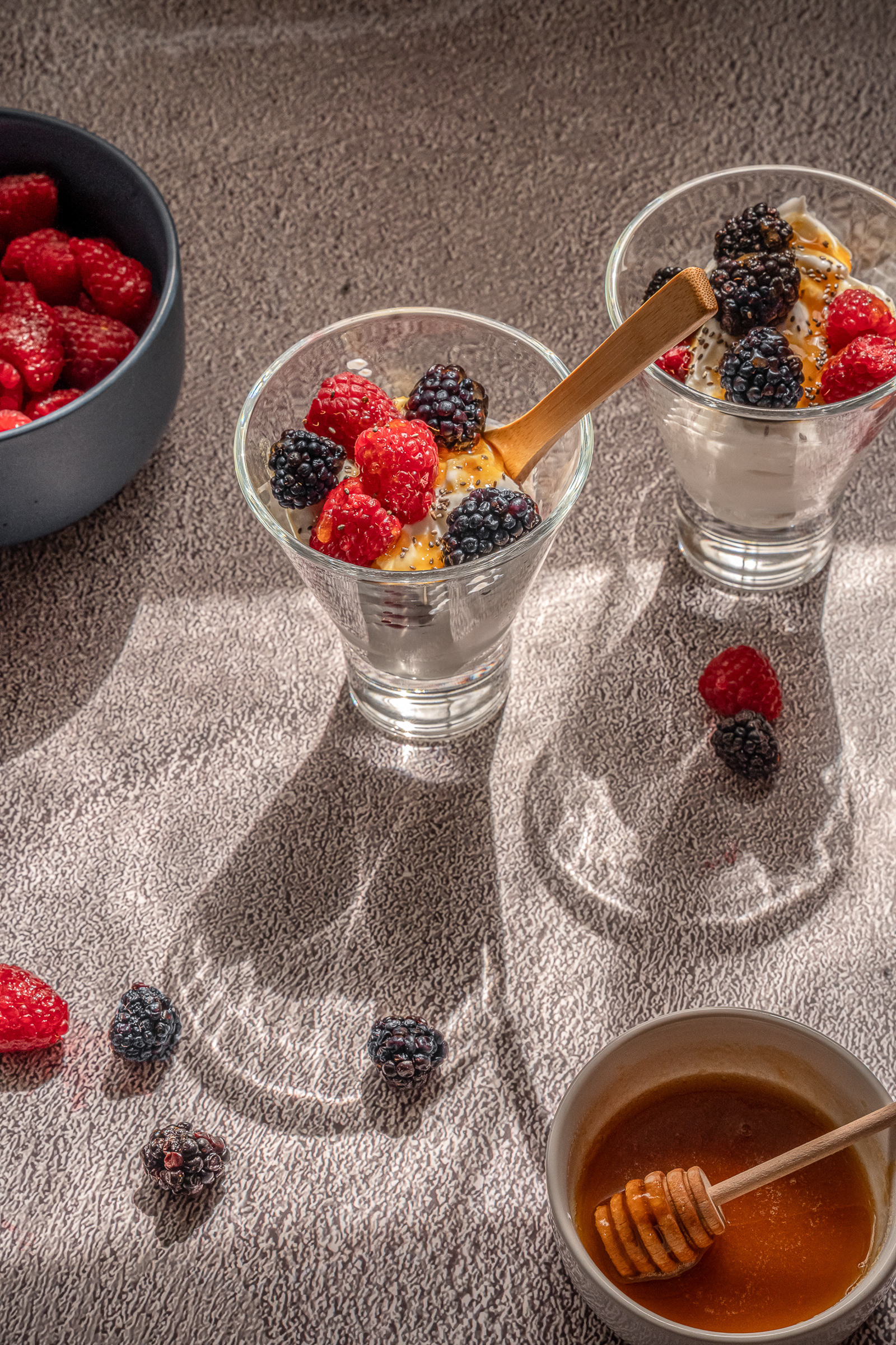 Side angle food photography of a yogurt berry parfait glass with a wooden spoon and a small bowl of honey, showing layers of fresh fruit