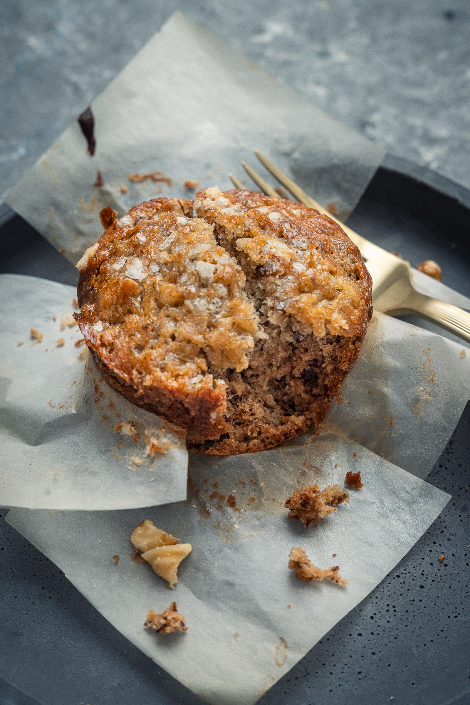 Macro food photography of a bitten banana walnut muffin on parchment paper with a gold dessert fork, showing a moist interior crumb and golden crust.