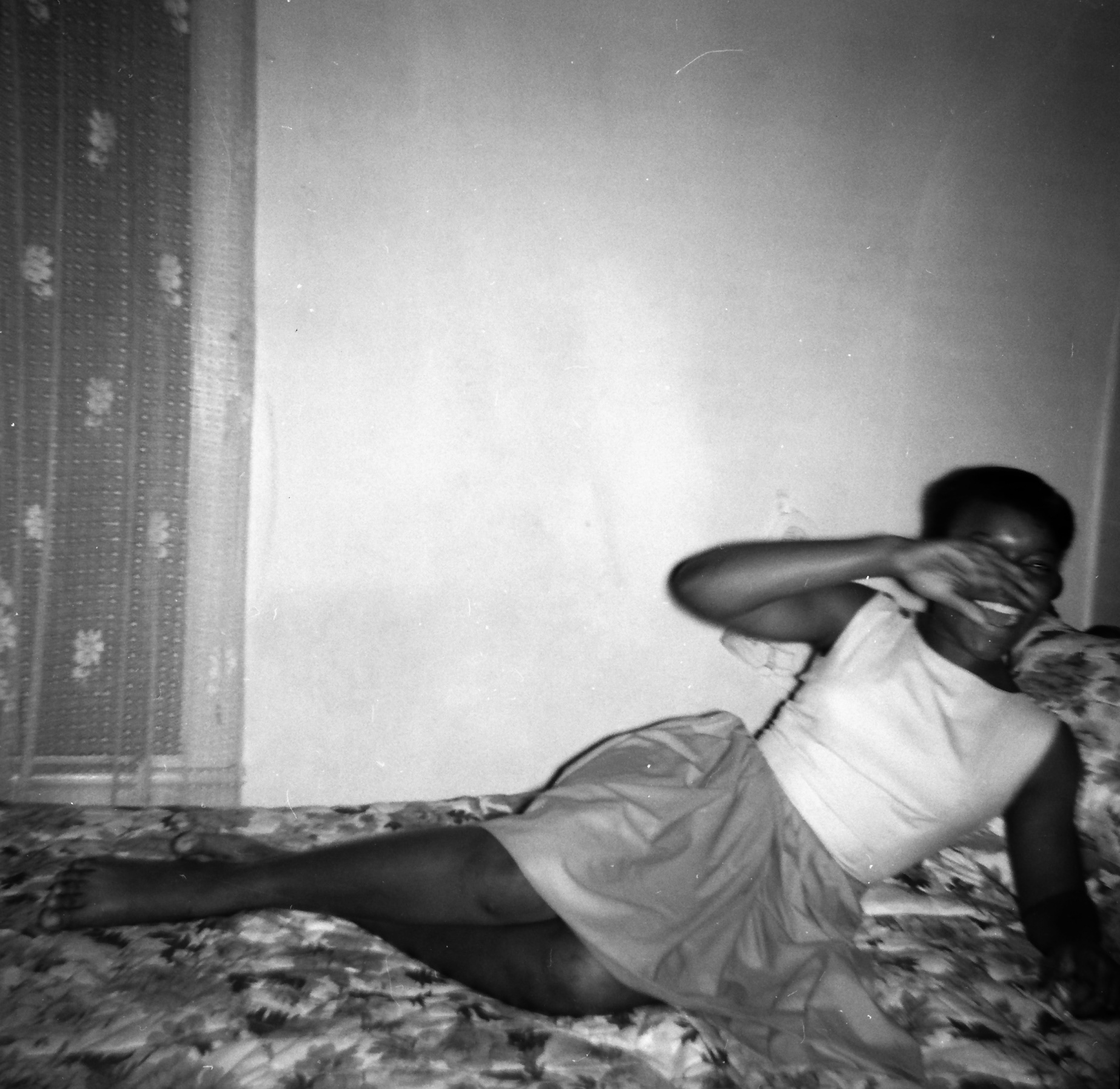 Black and white image of a Black teenager laughing on her bed with her hand covering her face. 