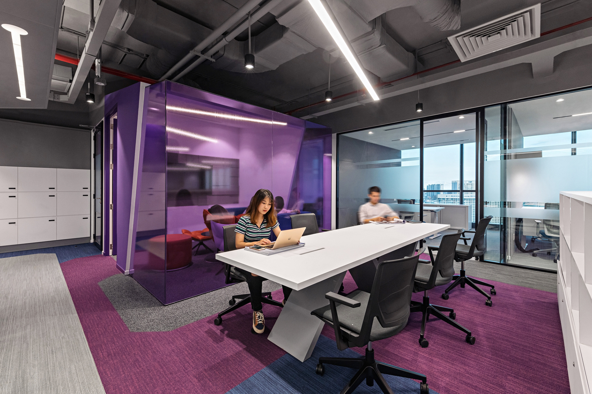 Two employees sitting at a white collaboration table in an open office space. There is purple, blue, and gray carpet beneath them and task chairs surrounding the table.