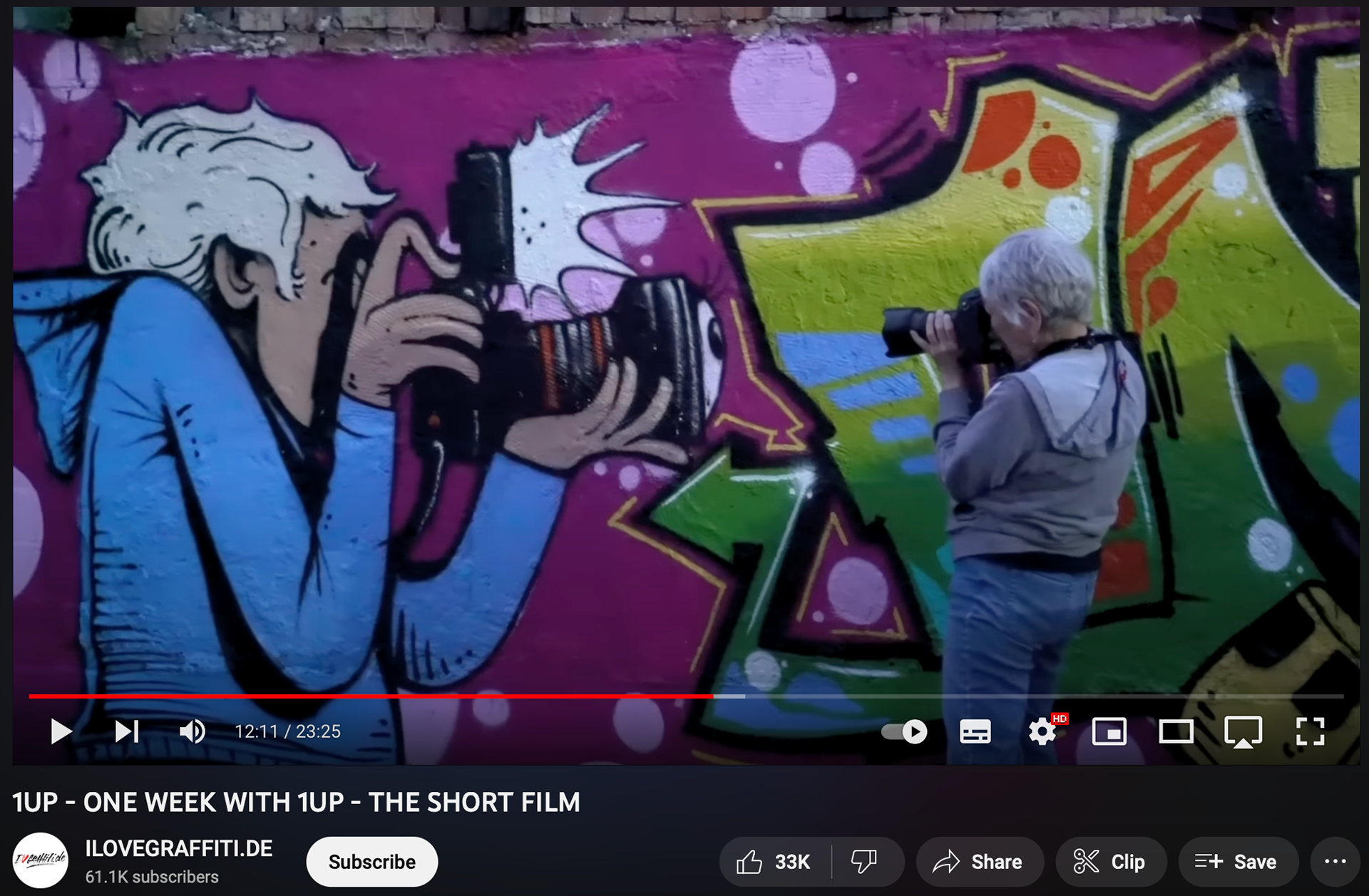 SCREENSHOT FROM VIDEO. MARTHA COOPER TAKING A PICTURE OF THE MURAL MADE BY THE 1 UP CREW.
