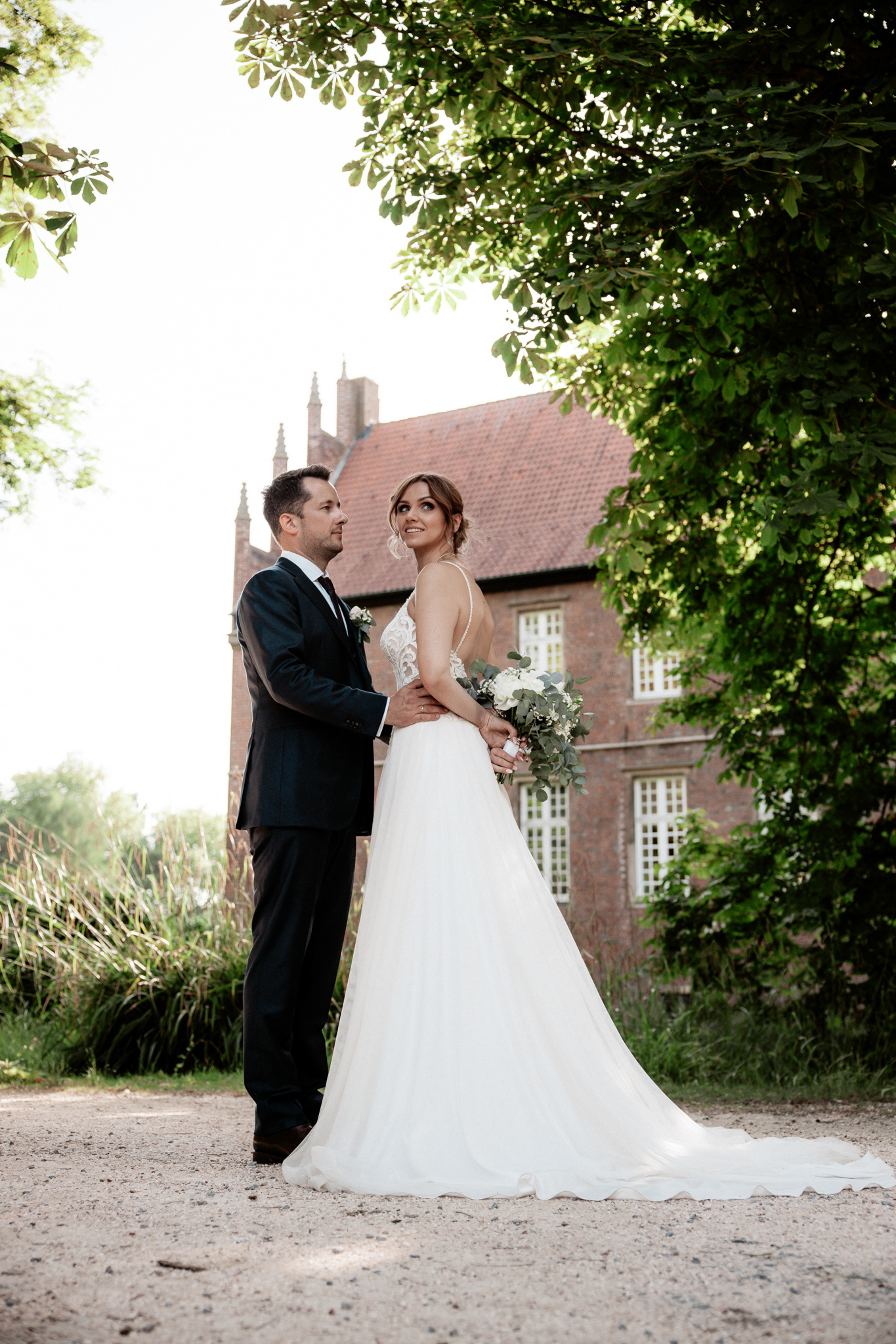 Braut Christina und Bräutigam Phillip in einem liebevollen Moment vor dem Schloss Herten. Die natürliche Umgebung und das sanfte Licht verleihen diesem Hochzeitsporträt eine märchenhafte Atmosphäre, während das Brautkleid und der Brautstrauß im Vordergrund stehen.