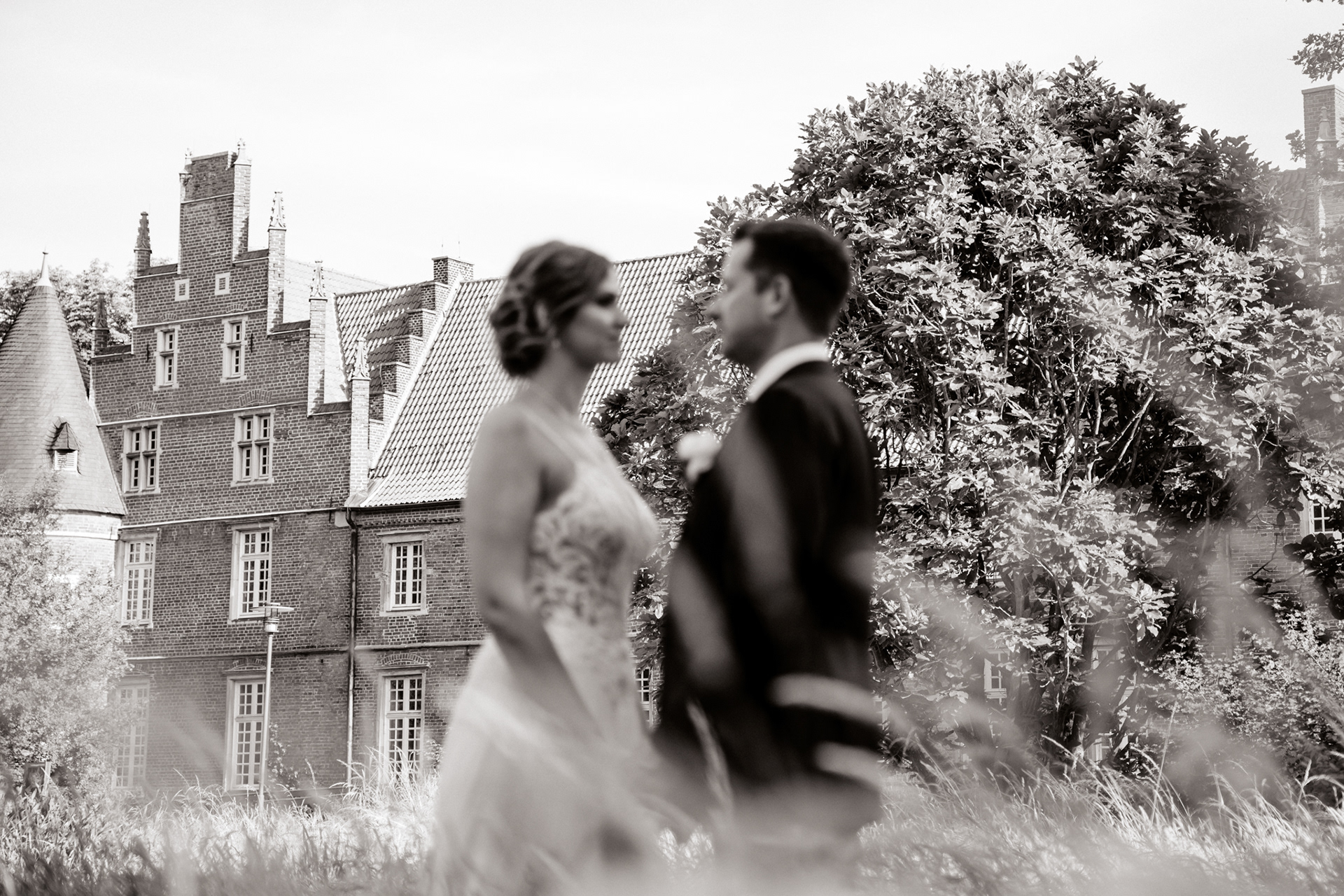 Romantisches Hochzeitsporträt von Christina & Phillip vor der historischen Kulisse des Schlosses Herten. Die unscharfe Darstellung des Brautpaares im Vordergrund sorgt für einen künstlerischen Effekt, während das Schloss mit seiner beeindruckenden Architektur in den Fokus rückt