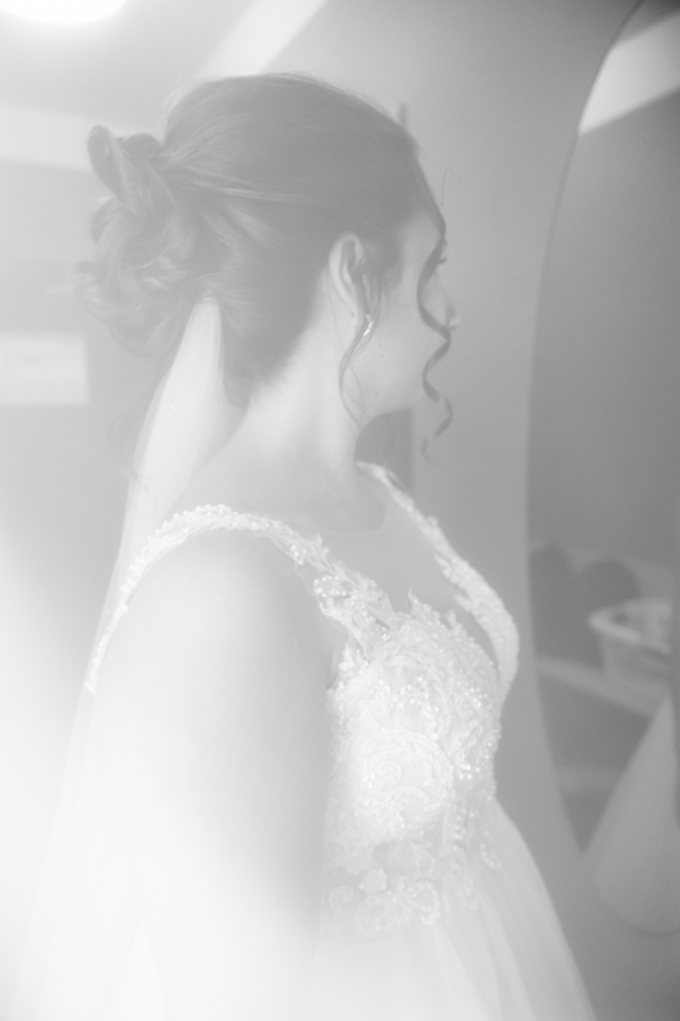 Künstlerisches Schwarz-Weiß-Foto von Braut Denise während des Getting Ready. Ihr Hochzeitskleid mit filigranen Spitzen- und Perlenverzierungen ist im sanften Licht zu sehen, während sie vor einem Spiegel steht. Der unscharfe, verträumte Effekt verleiht dem Bild eine emotionale Tiefe.