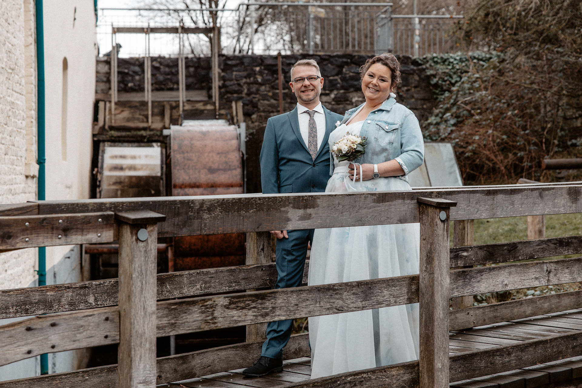 Das frisch verheiratete Paar steht lächelnd auf einer Holzbrücke vor dem Wasserrad der Goldberger Mühle in Mettmann.