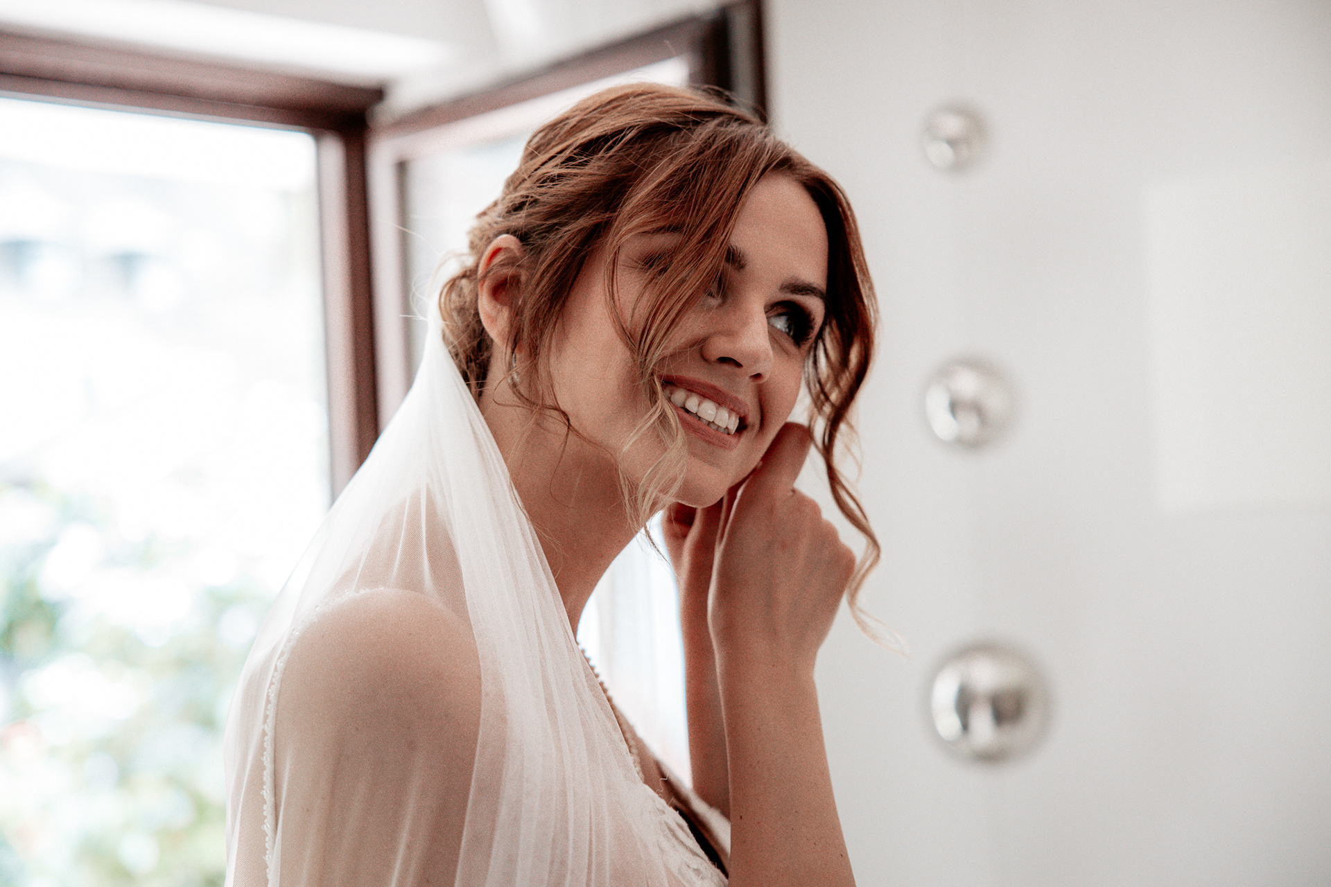 Christina beim Getting Ready. Die Braut steckt sich ihre Ohrringe an und lächelt voller Vorfreude auf ihren großen Tag. Hochzeitsfotografie mit natürlichen Momenten beim Gettin Ready.