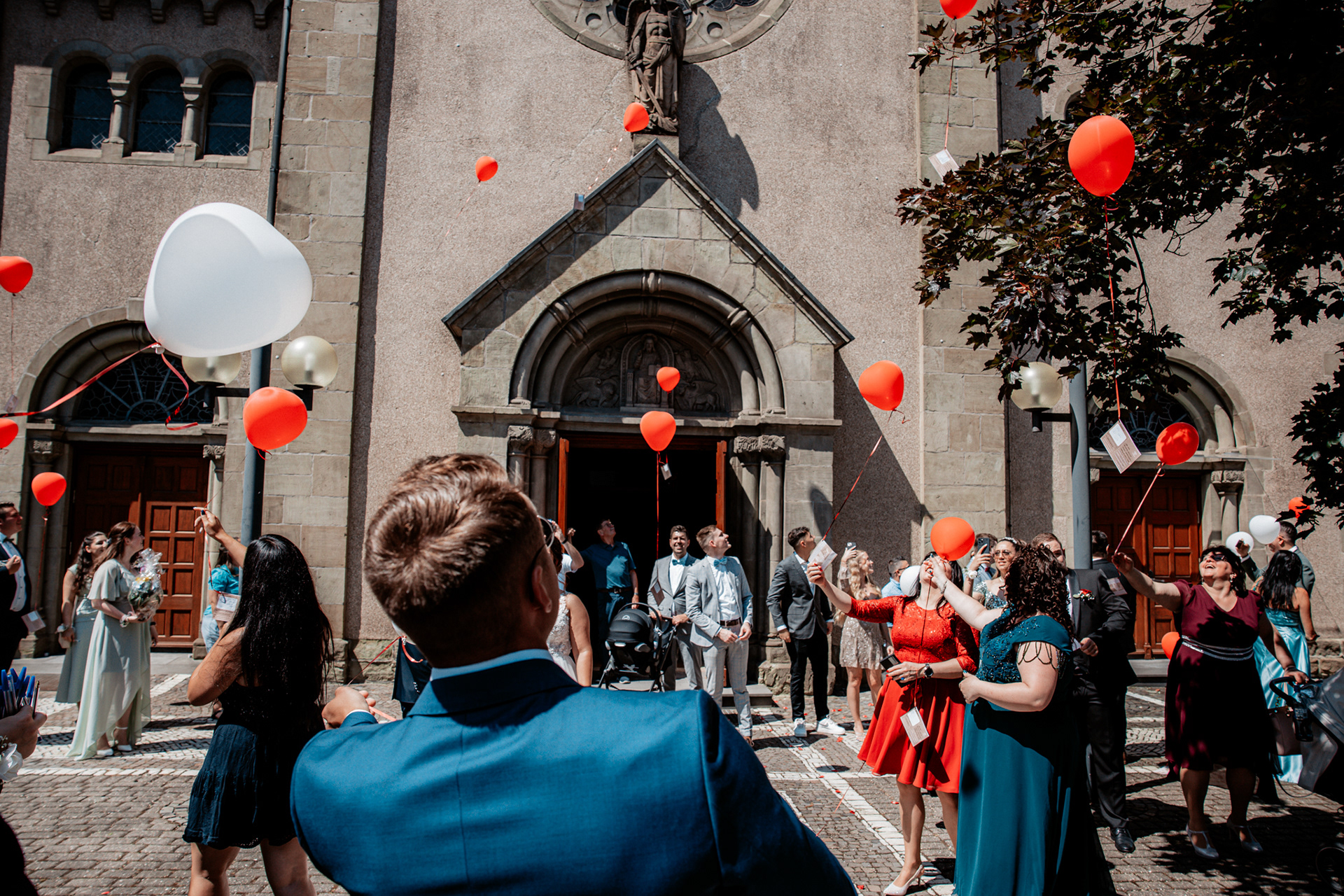 Die Hochzeitsgesellschaft lässt rote und weiße Luftballons in den Himmel steigen, während das Brautpaar vor der Kirche steht. Die Gäste feiern ausgelassen diesen emotionalen Moment.