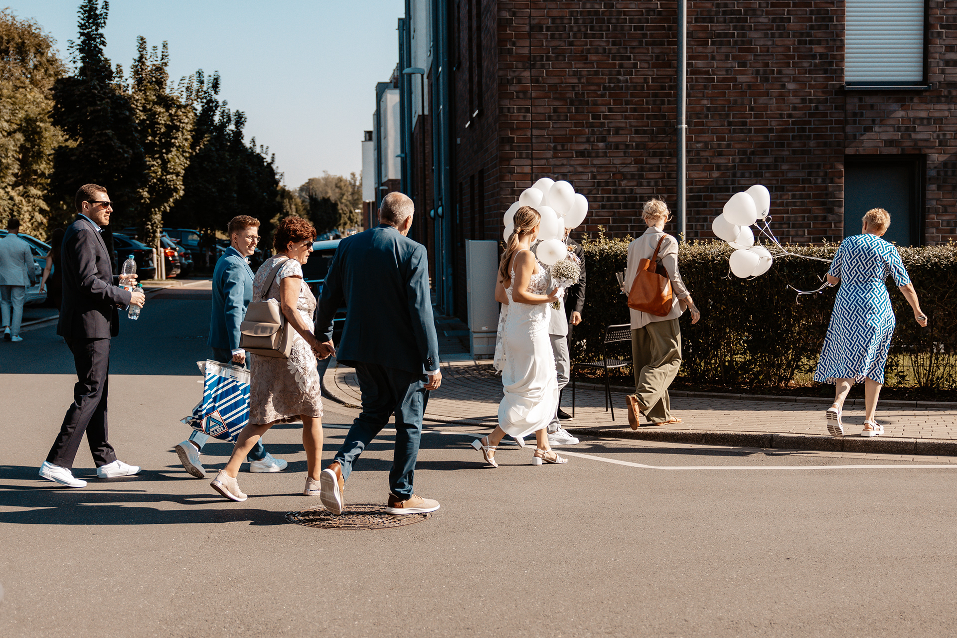 Hochzeitsgesellschaft auf dem Weg zur nächsten Location, Braut Justine hält ihren Brautstrauß und weiße Luftballons in der Hand.