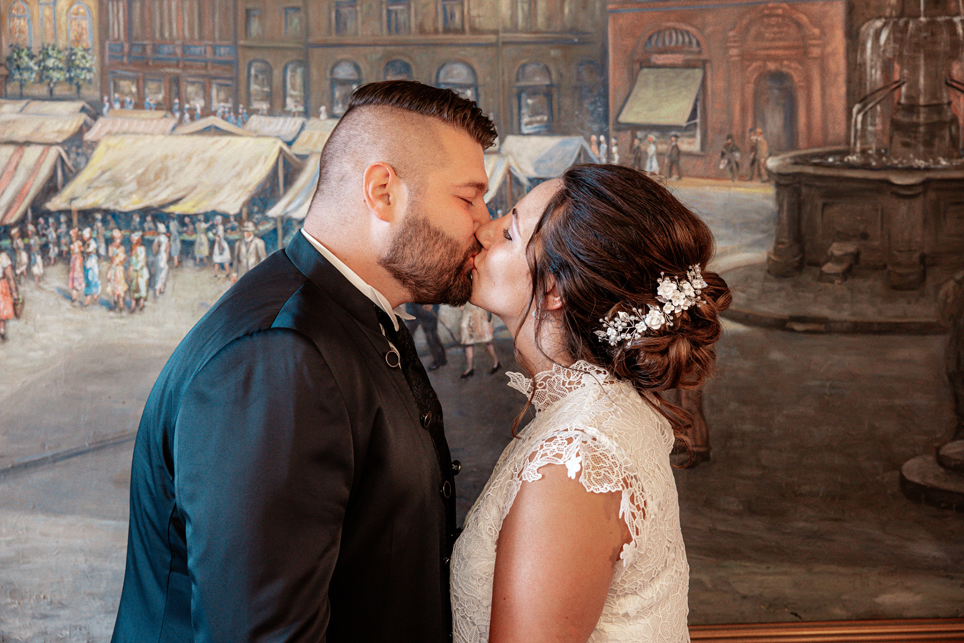 Denise und Michael küssen sich vor einem großen historischen Wandgemälde im Standesamt Dortmund. Die Braut trägt ein elegantes zweiteiliges Hochzeitskleid mit Spitze, während der Bräutigam in einem dunklen Anzug mit Weste gekleidet ist. Ein festlicher Moment ihrer standesamtlichen Trauung.