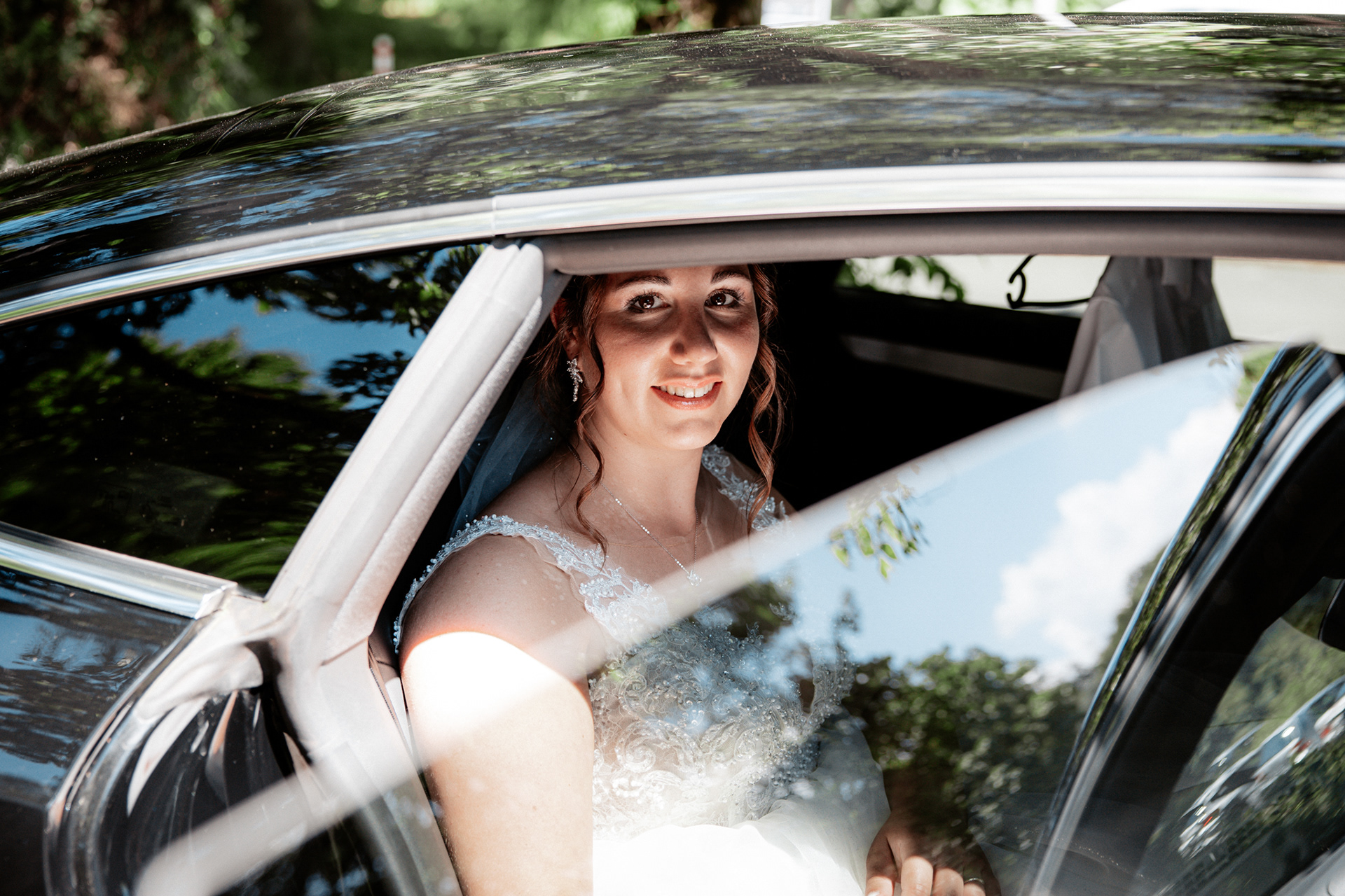 Strahlende Braut Denise sitzt im Hochzeitsauto und blickt voller Vorfreude aus dem Fenster. Das Sonnenlicht spiegelt sich auf dem glänzenden schwarzen Wagen, während sie sich auf die Fahrt zur Trauung vorbereitet.