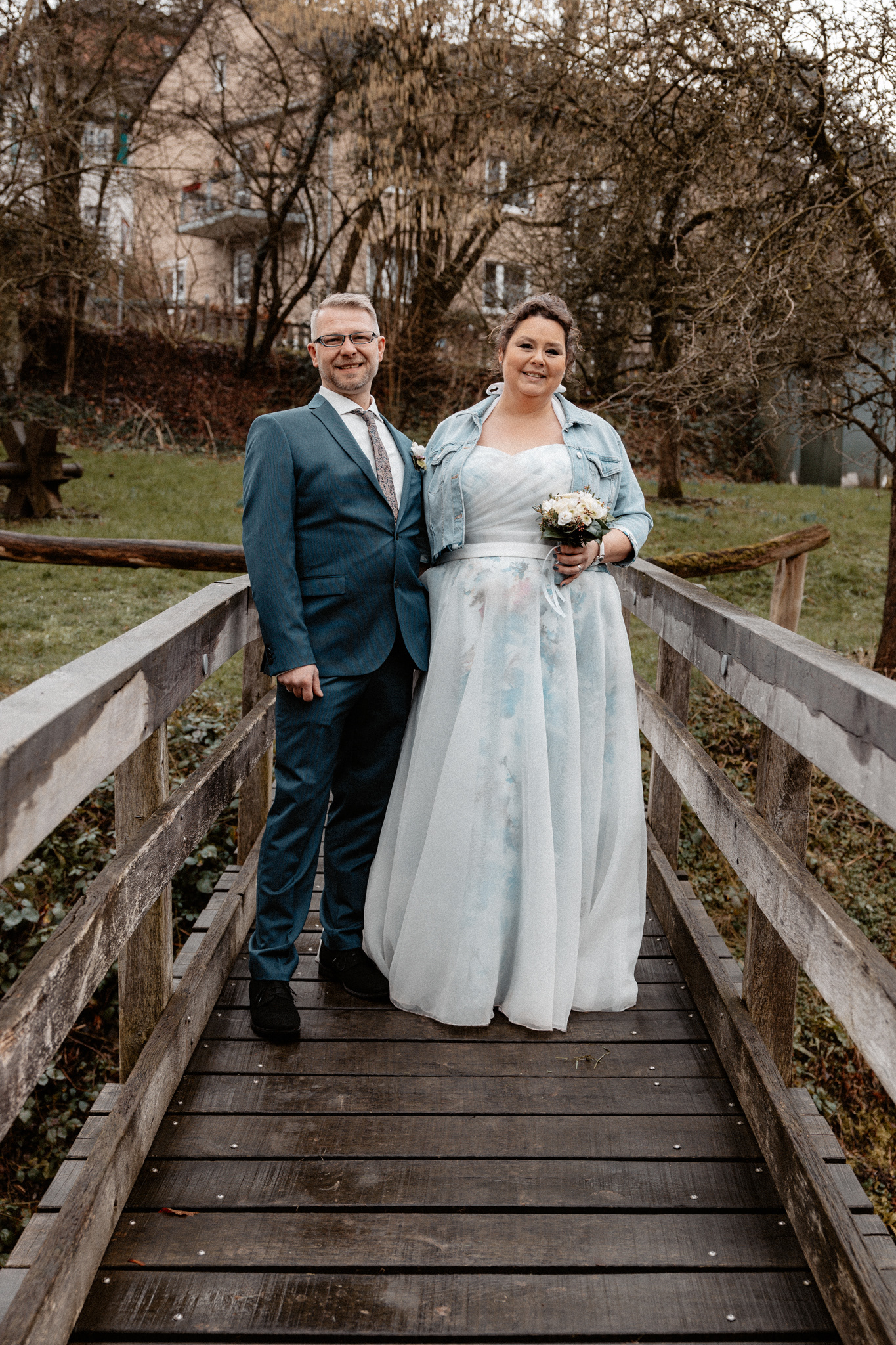 Claudia und Sven als frisch verheiratetes Paar auf einer idyllischen Holzbrücke in Mettmann. Die Braut trägt ein fließendes, individuelles Hochzeitskleid mit blauen Akzenten und eine Jeansjacke, während der Bräutigam stilvoll im blauen Hochzeitsanzug posiert. Hochzeitsfotos NRW.