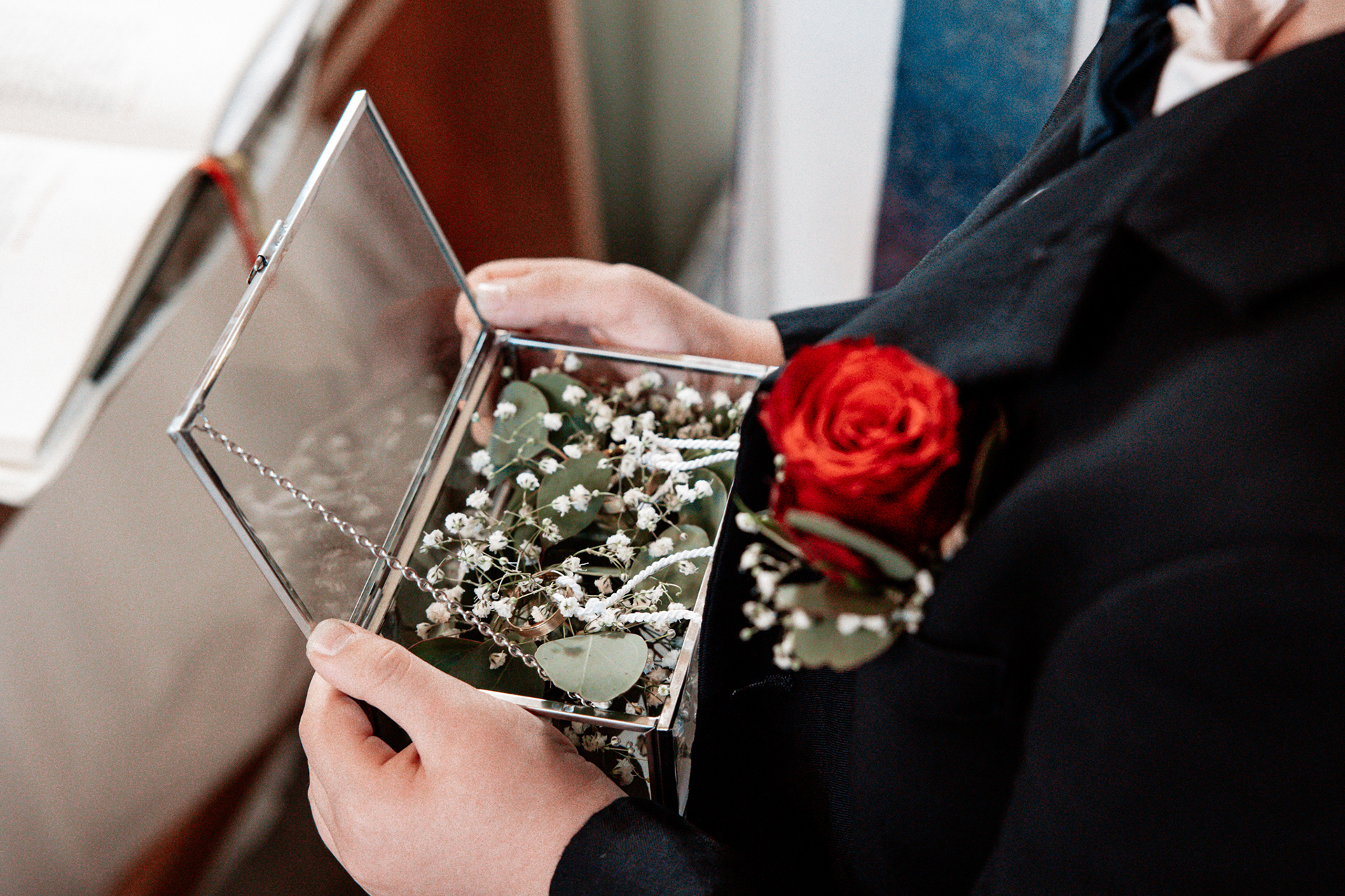 Der Ringträger hält eine elegante Glasbox mit den Eheringen von Denise und Michael. Die Ringe liegen auf einem Bett aus Eukalyptusblättern und Schleierkraut, während eine rote Rose die romantische Szene abrundet.