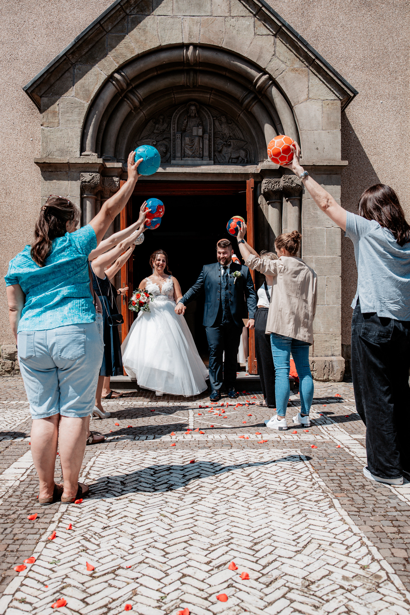 Denise und Michael verlassen strahlend die Kirche als frisch vermähltes Ehepaar. Freunde und Familie bilden ein Spalier und halten bunte Handbälle in die Luft, während rote Rosenblätter auf dem Boden verstreut sind.