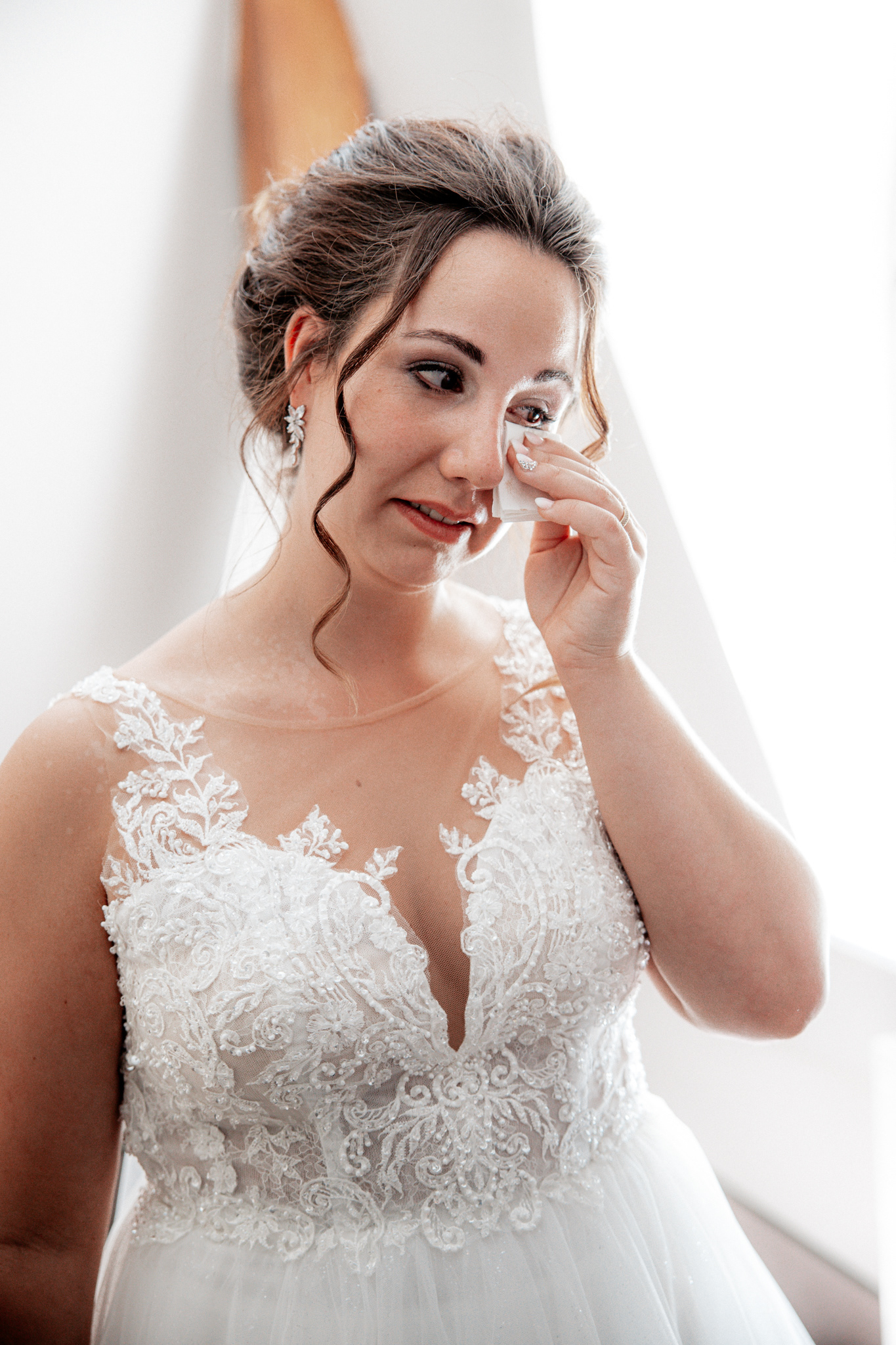 Emotionale Braut Denise wischt sich eine Träne aus dem Gesicht während des Getting Ready. Sie trägt ein elegantes, weißes Hochzeitskleid mit feinen Spitzen- und Perlenverzierungen. Das natürliche Licht aus dem Fenster verstärkt die emotionale Stimmung dieses besonderen Moments vor der Trauung