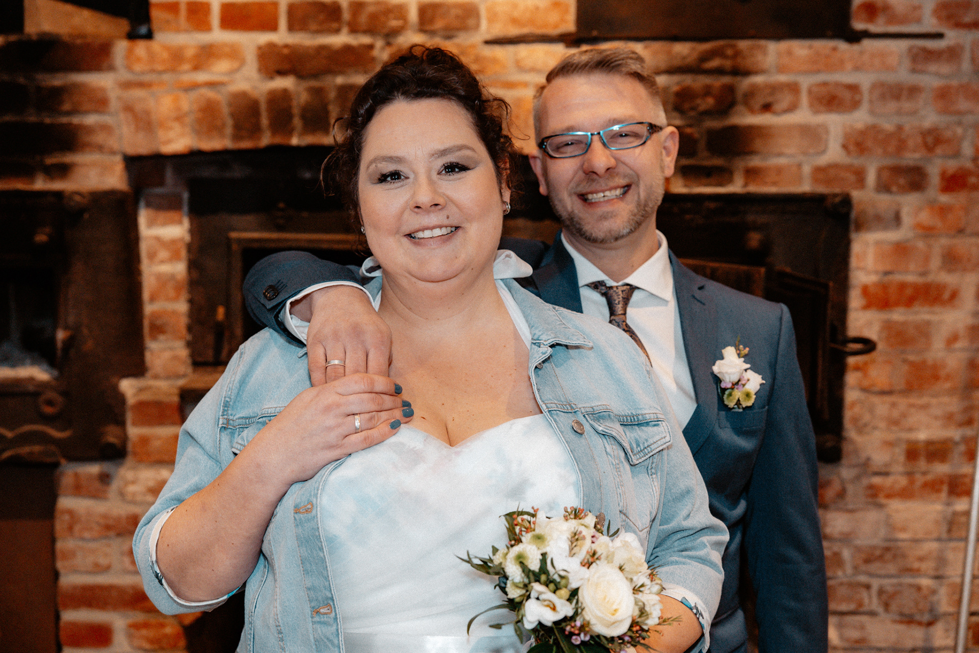 Strahlendes Brautpaar Claudia und Sven vor einer rustikalen Backsteinwand in der Goldberger Mühle, Mettmann. Die Braut trägt ein modernes Hochzeitskleid mit Jeansjacke, während der Bräutigam im eleganten blauen Anzug lächelt. Hochzeitsfotografie NRW