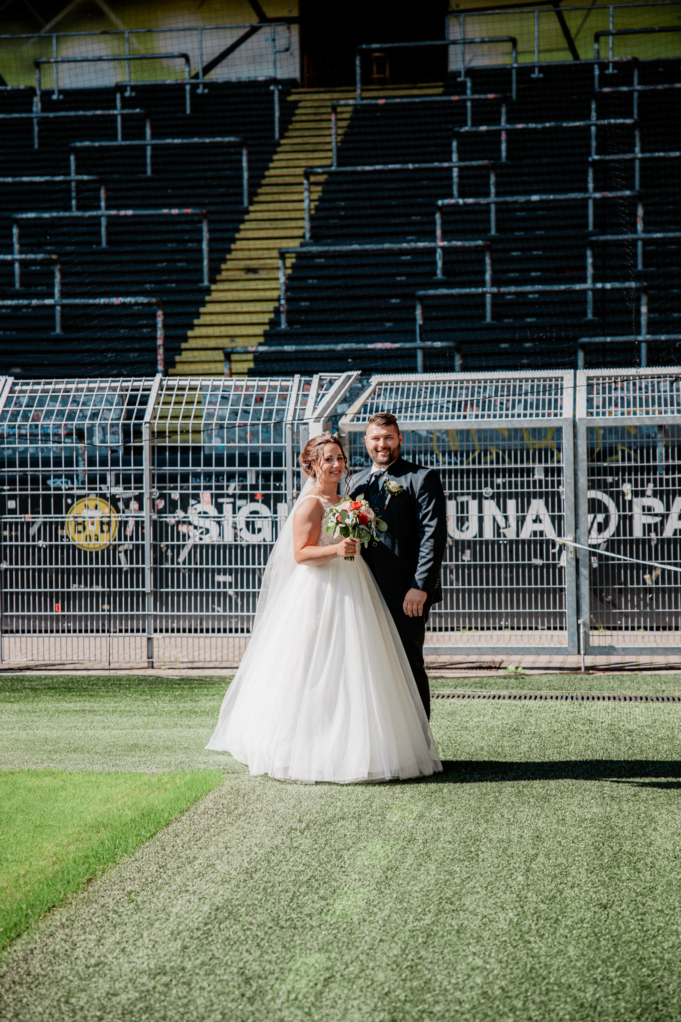 Hochzeitsfoto von Denise und Michael im Signal Iduna Park in Dortmund. Das Brautpaar posiert glücklich auf dem Spielfeld des berühmten Fußballstadions von Borussia Dortmund, umgeben von der beeindruckenden Kulisse der leeren Tribünen.