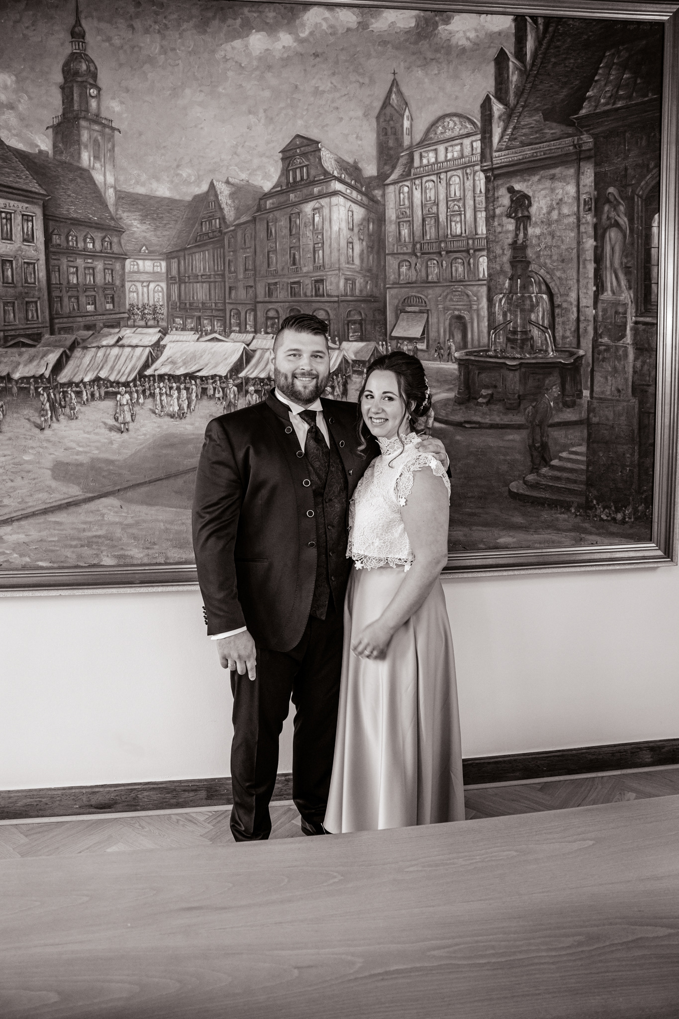 Denise und Michael stehen strahlend vor einem großen historischen Wandgemälde im Standesamt Dortmund. Die Braut trägt ein elegantes zweiteiliges Hochzeitskleid mit Spitze, während der Bräutigam in einem dunklen Anzug mit Weste gekleidet ist. Ein festlicher Moment ihrer standesamtlichen Trauung.