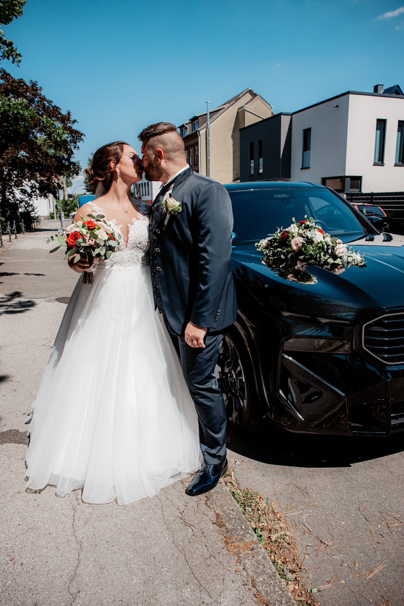 Romantischer Hochzeitsmoment: Denise und Michael küssen sich innig vor ihrem eleganten schwarzen Hochzeitsauto. Die Braut hält einen stilvollen Strauß mit roten und weißen Rosen, während das Auto ebenfalls mit Blumen geschmückt ist.