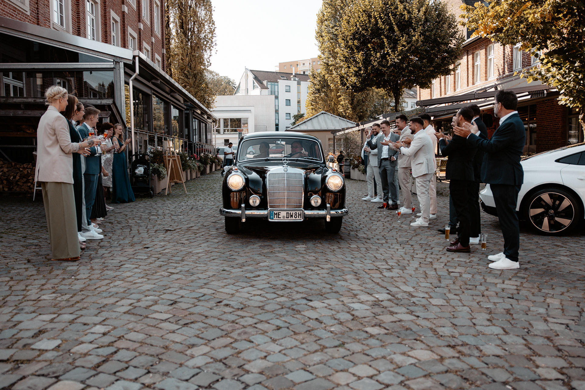Ein klassischer schwarzer Mercedes Oldtimer fährt durch eine gepflasterte Straße, flankiert von applaudierenden Hochzeitsgästen.