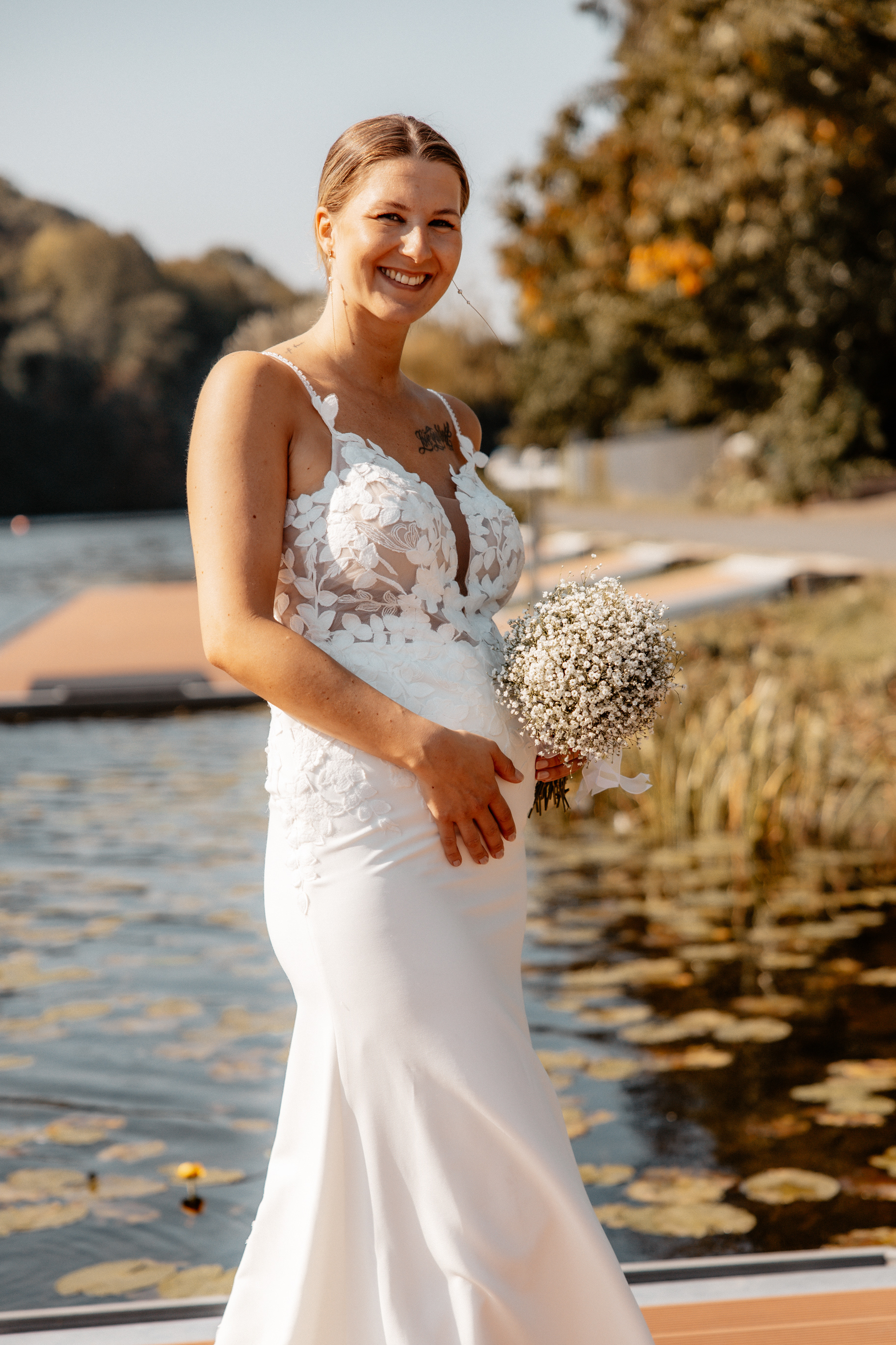 Die Braut Justine posiert lächelnd mit ihrem Brautstrauß am Wasser, das Licht reflektiert sich sanft auf ihrer Haut.
